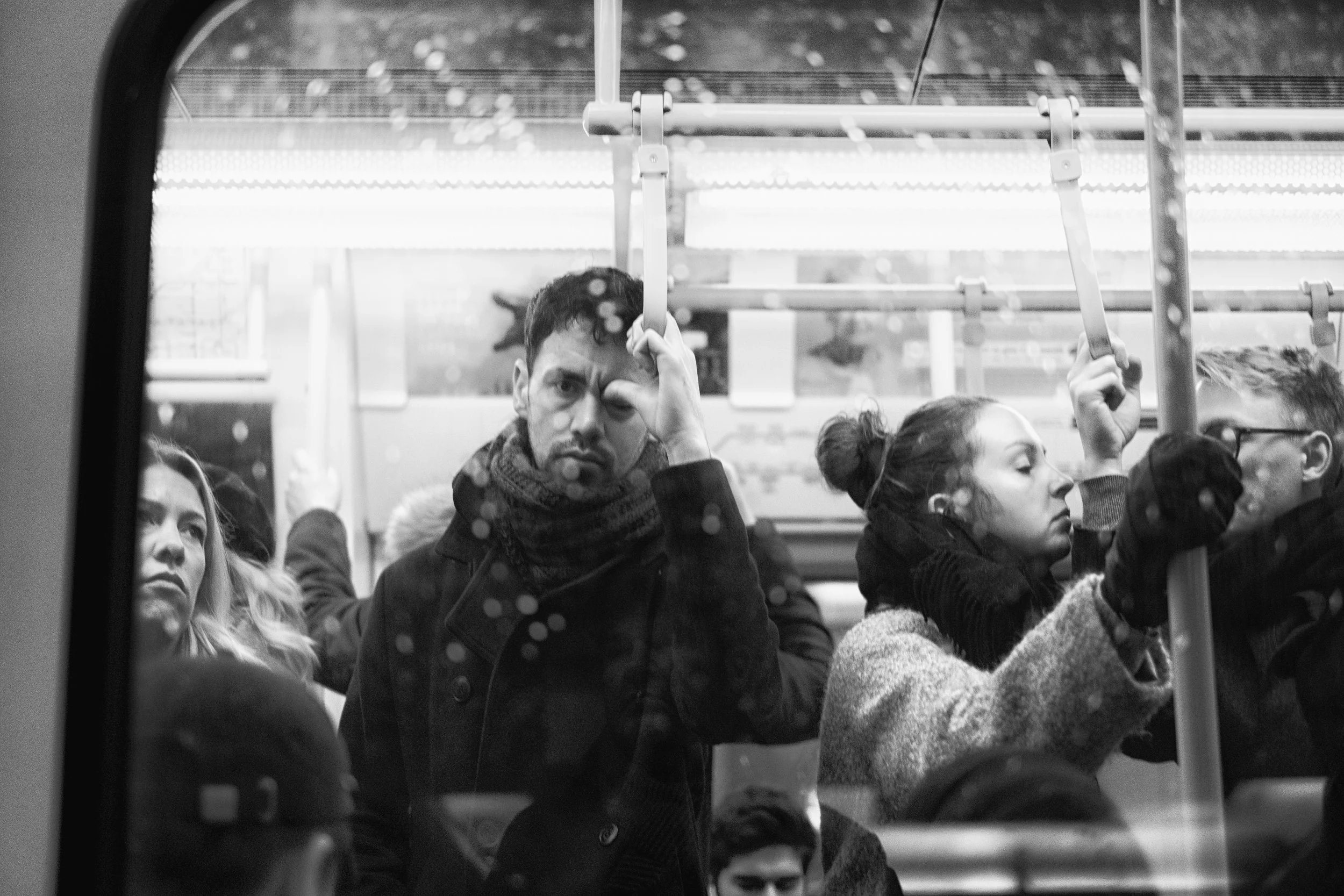 Black and white photo of people standing inside a bus. A man in a jacket and scarf looks directly at the camera with a somewhat serious expression, holding on to a bus handrail. To his right, a woman with her hair tied up, wearing a coat, is looking 