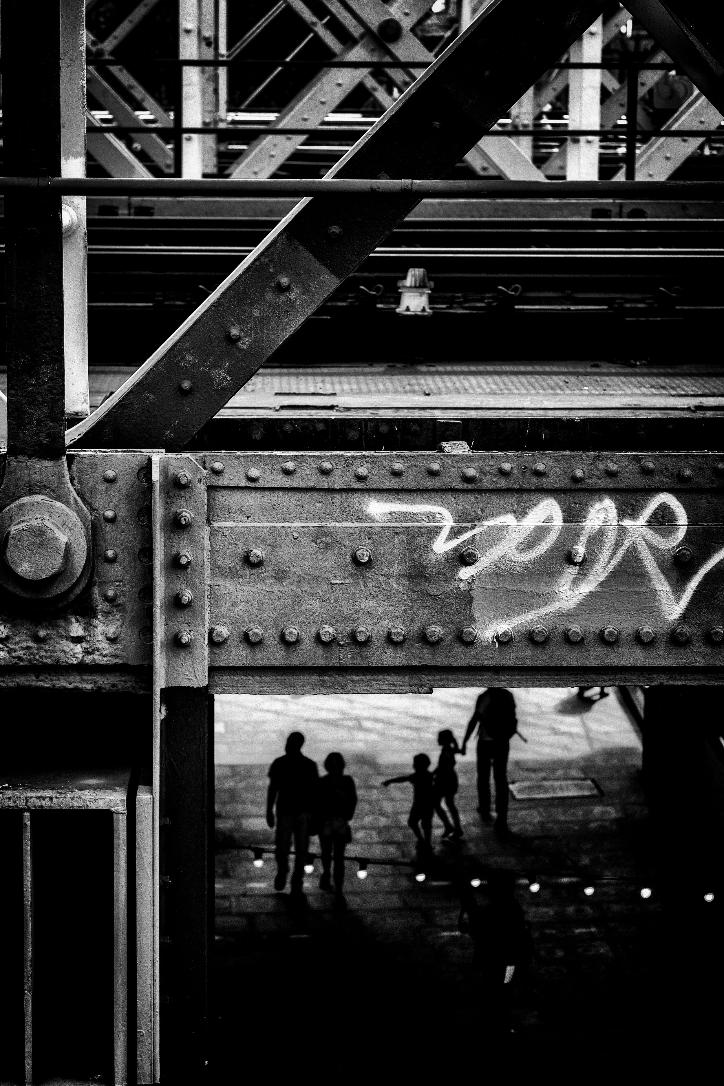 Black and white photo of people walking under a large metal bridge with rivets and beams, with graffiti on the bridge