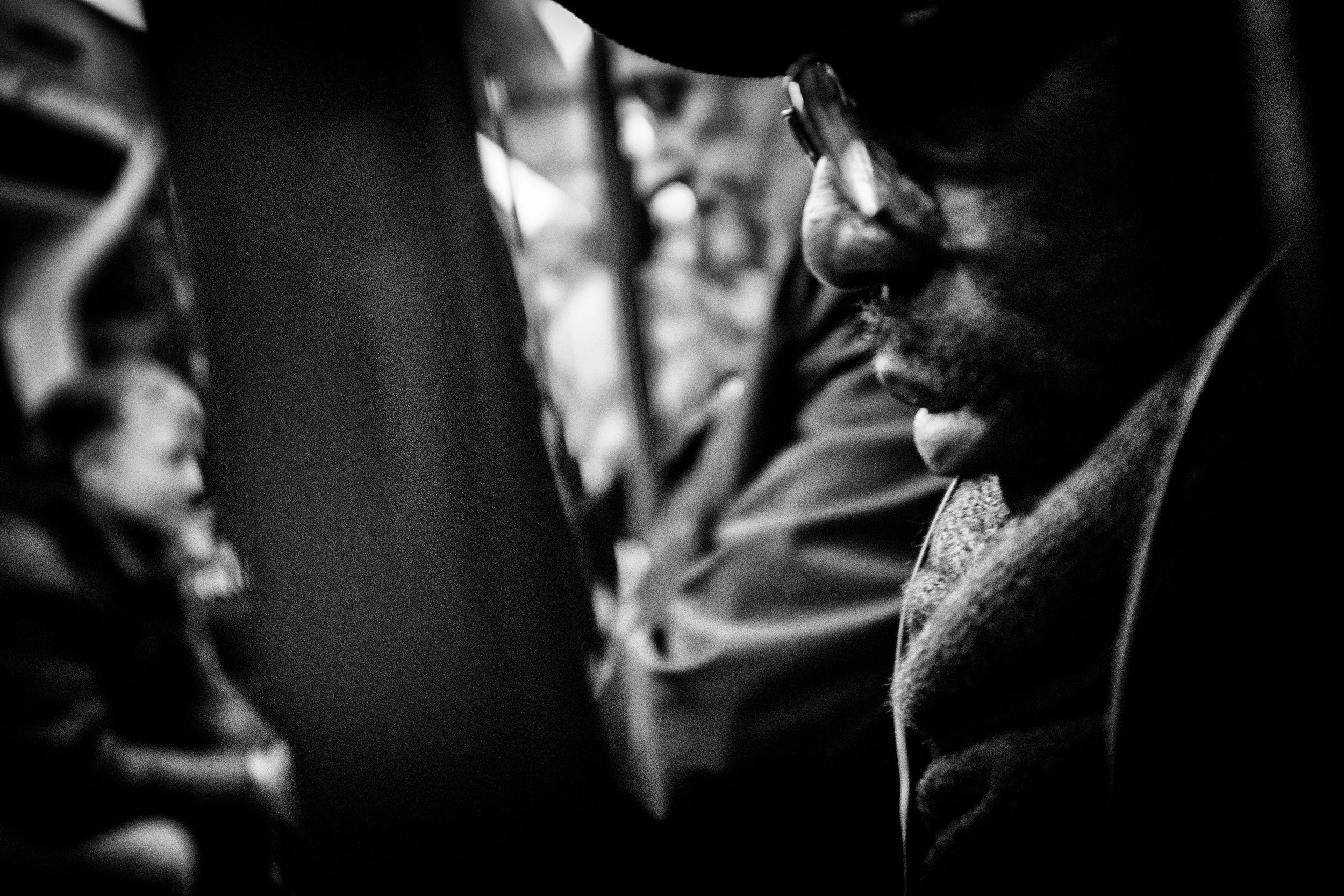 Close-up of a man wearing glasses in profile, with blurred background including a woman, in black and white.