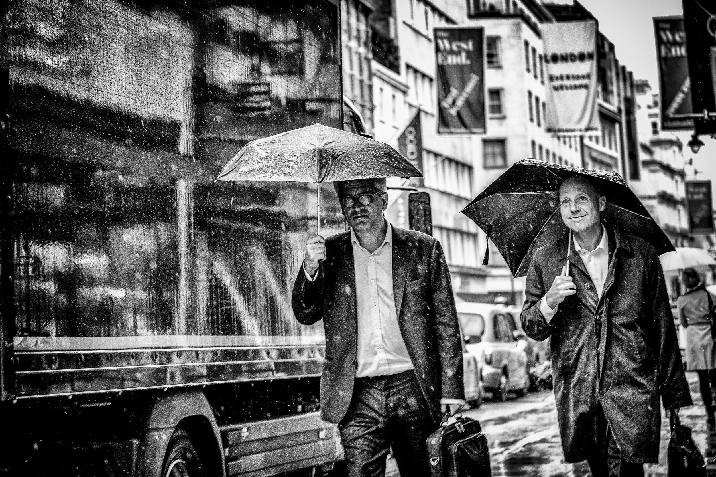 Two men walking down a city street in rain, holding umbrellas; one has glasses and carries a briefcase, both are wearing suits and coats, with storefronts and signs visible in the background.