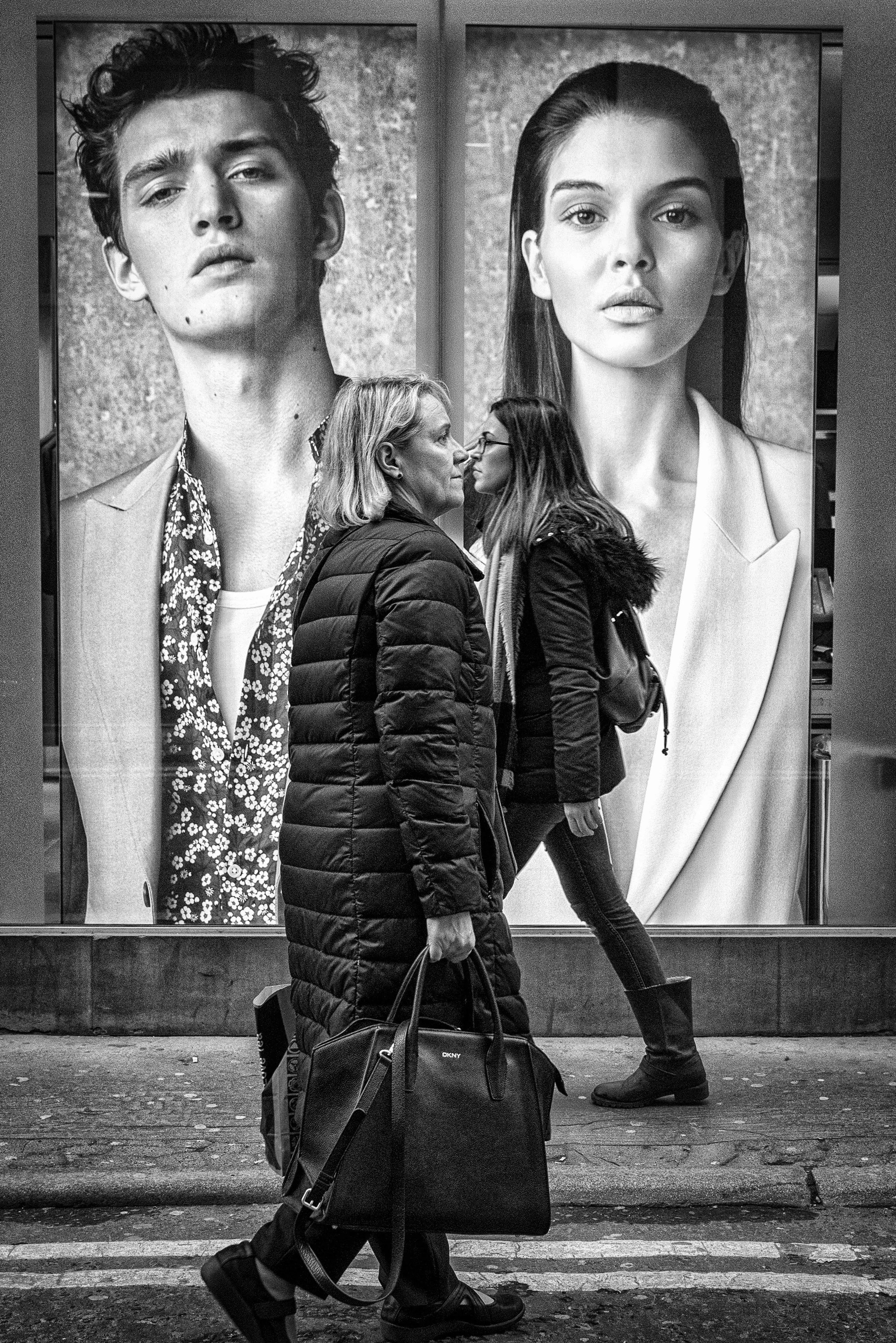 Two women dressed in winter clothing walking past a large advertisement featuring two large black-and-white portraits of a young man and a woman.