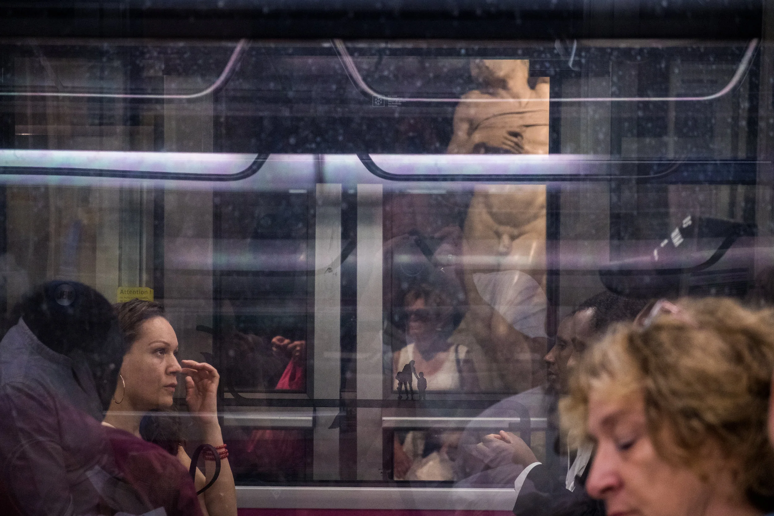 People sitting inside a train or subway, seen through the window, with reflections of the platform and other passengers.