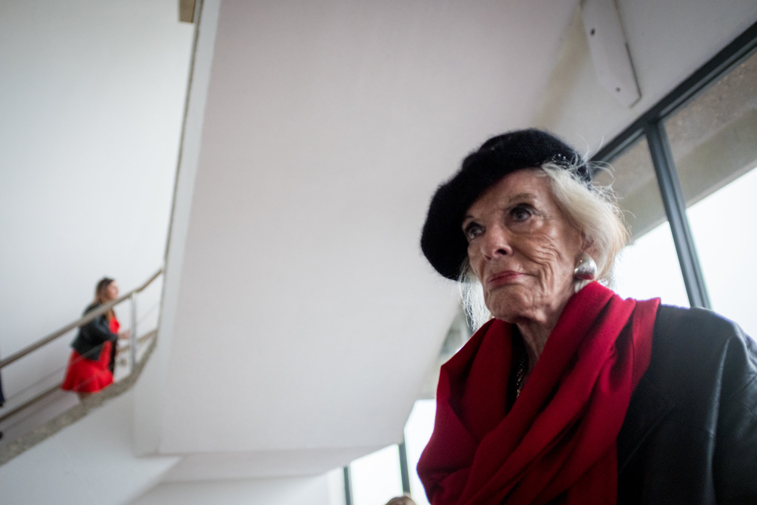 An elderly woman with blonde hair wearing a black hat, red scarf, and earrings sitting indoors near a window, with a woman in red and black in the background on a staircase.