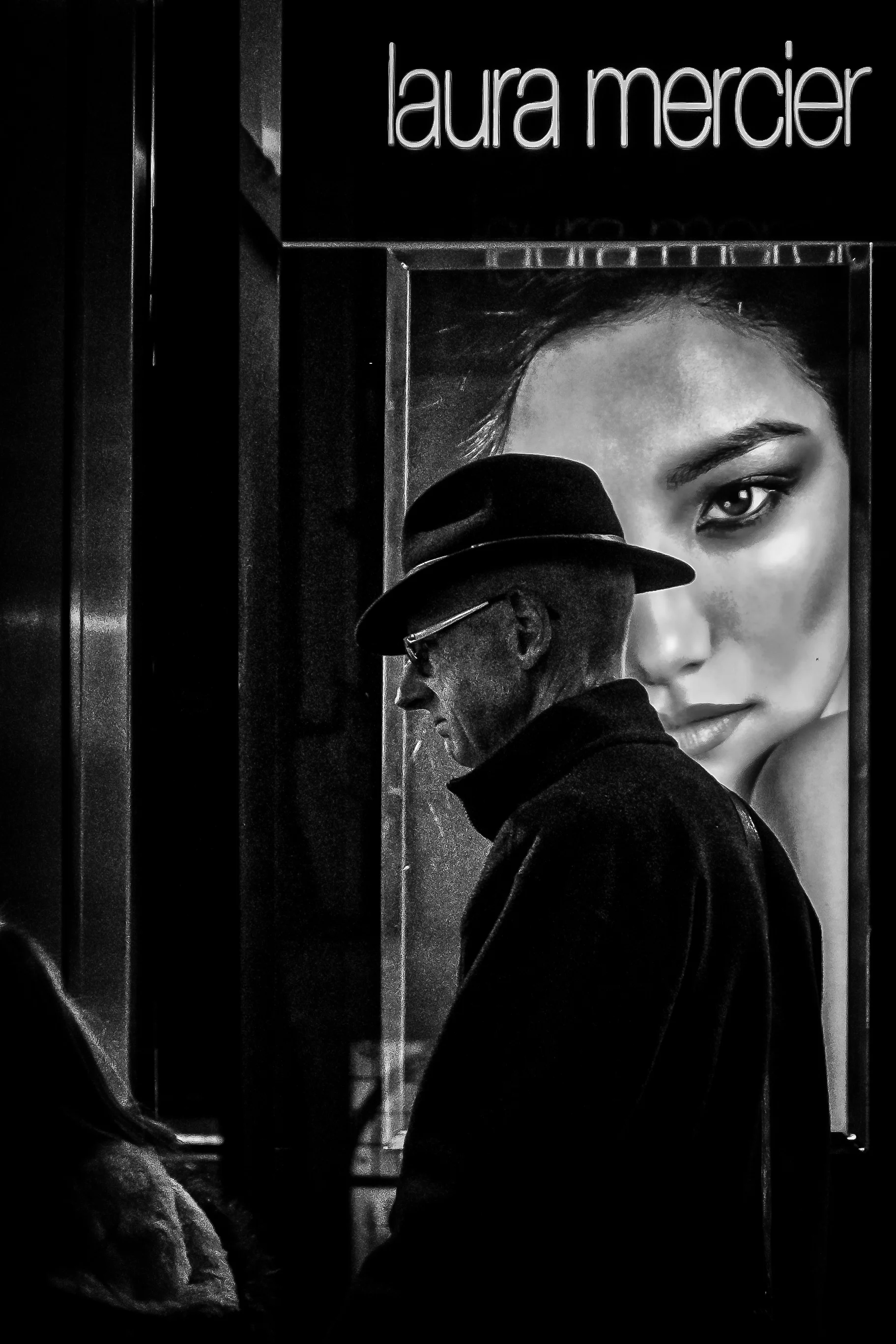 A black and white photo of an older man wearing a hat and glasses, looking down, standing in front of a large poster featuring a woman's face.