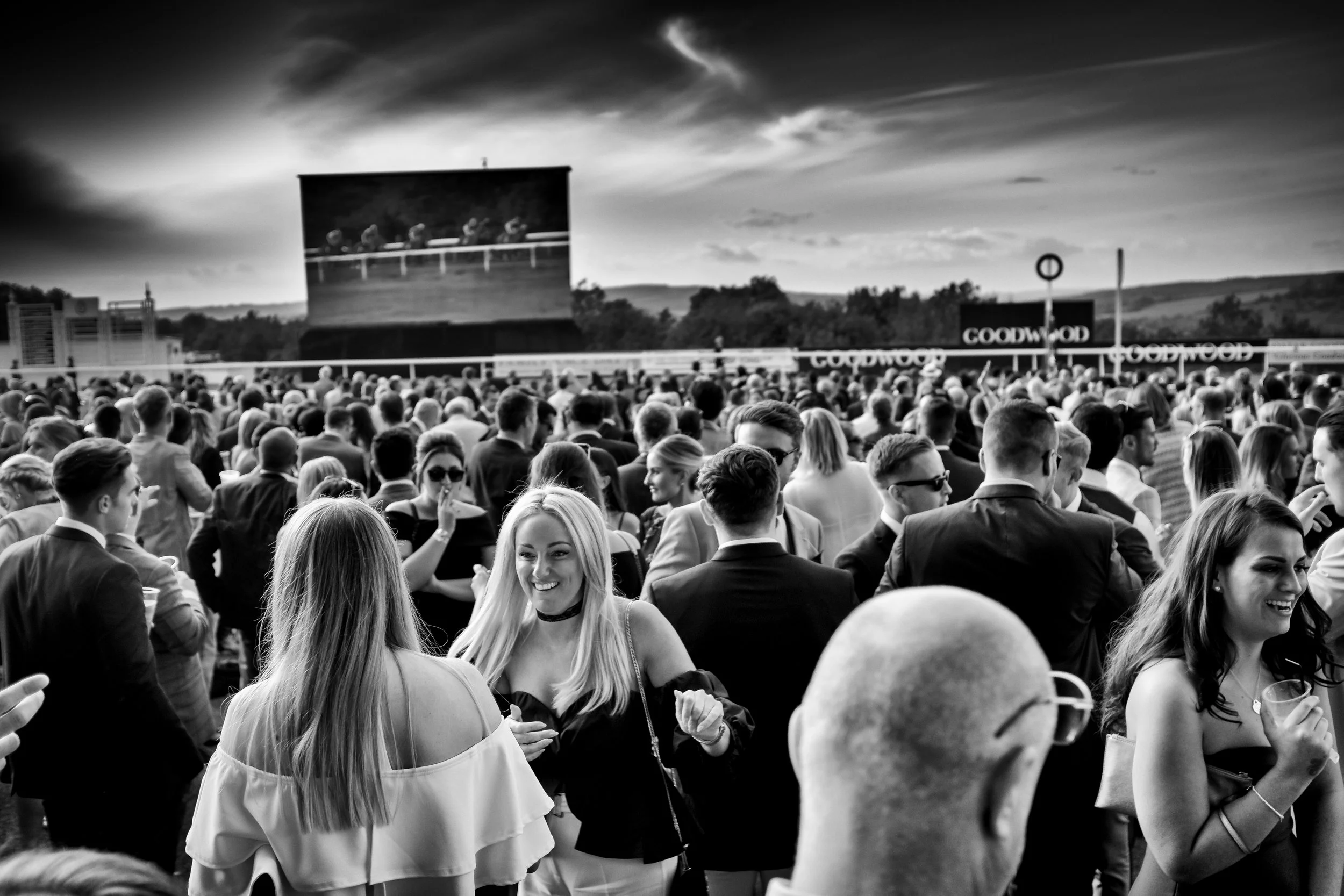 A black and white photo of a crowd of people at an outdoor event, with some wearing sunglasses and engaged in conversation, facing a large screen displaying a horse race, under a sky with clouds.