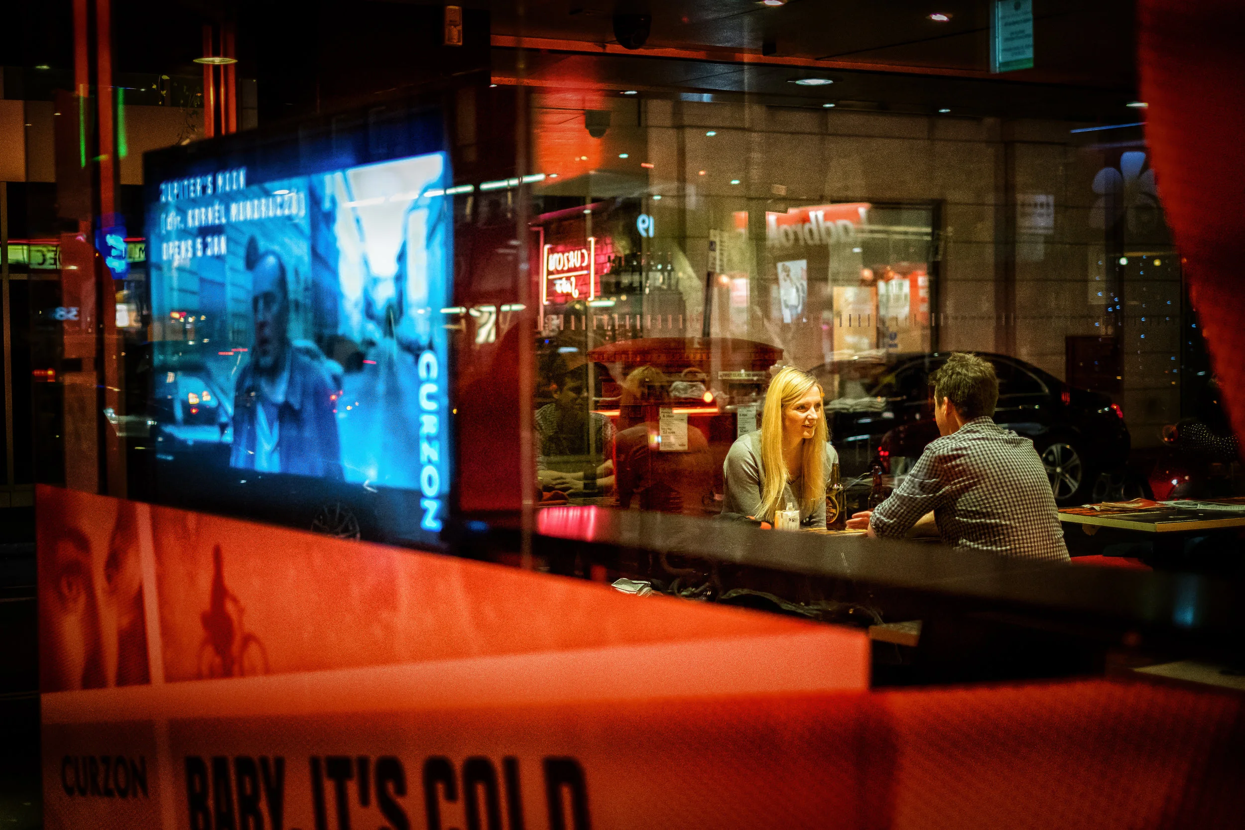 Two people sitting at a table inside a restaurant, seen through a window at night, with city lights and signs visible outside.