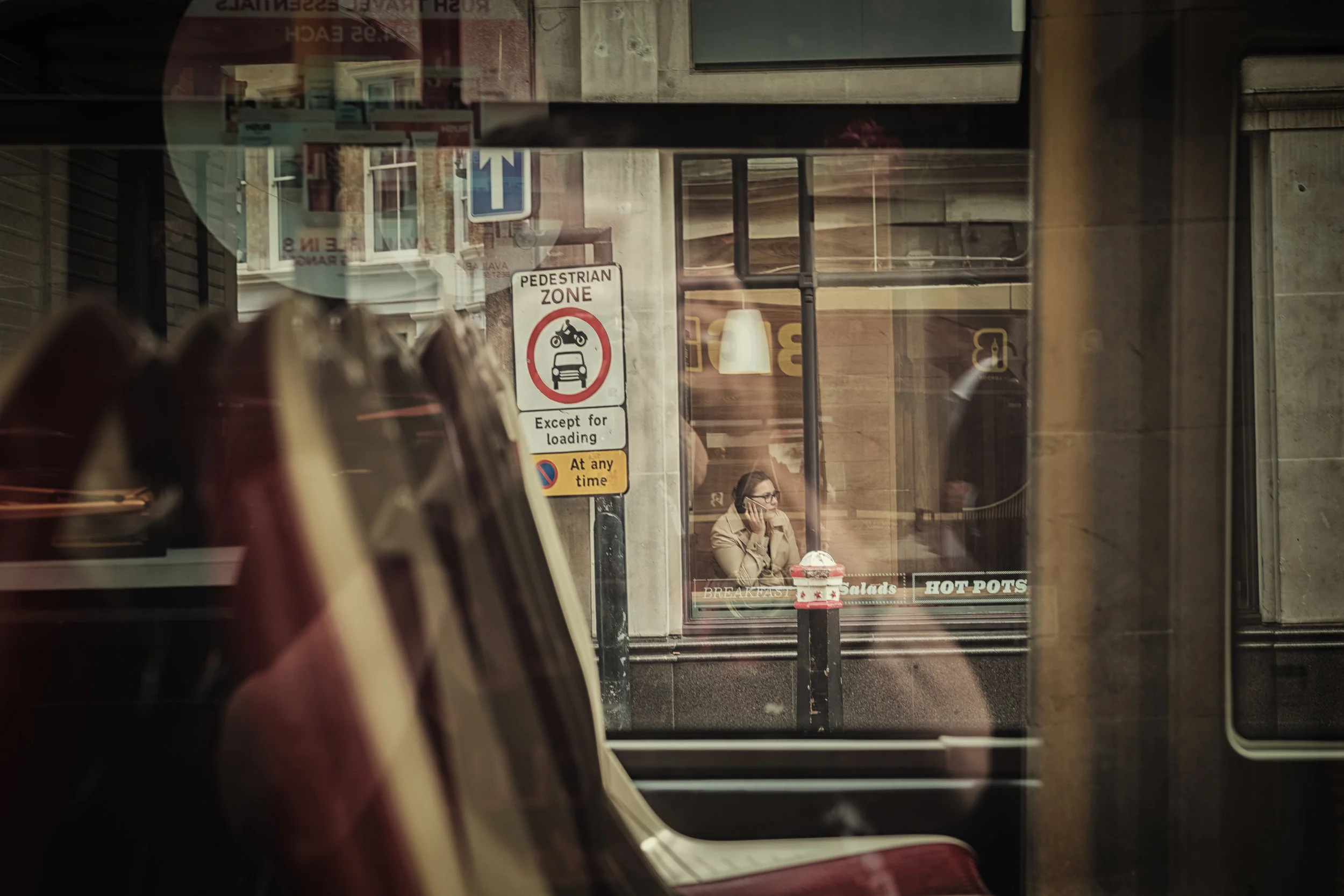 Inside a bus, view through the window shows a woman with glasses sitting at a table outside a café, talking on her phone. The café has a sign advertising breakfast, salads, hot pots, and other items.