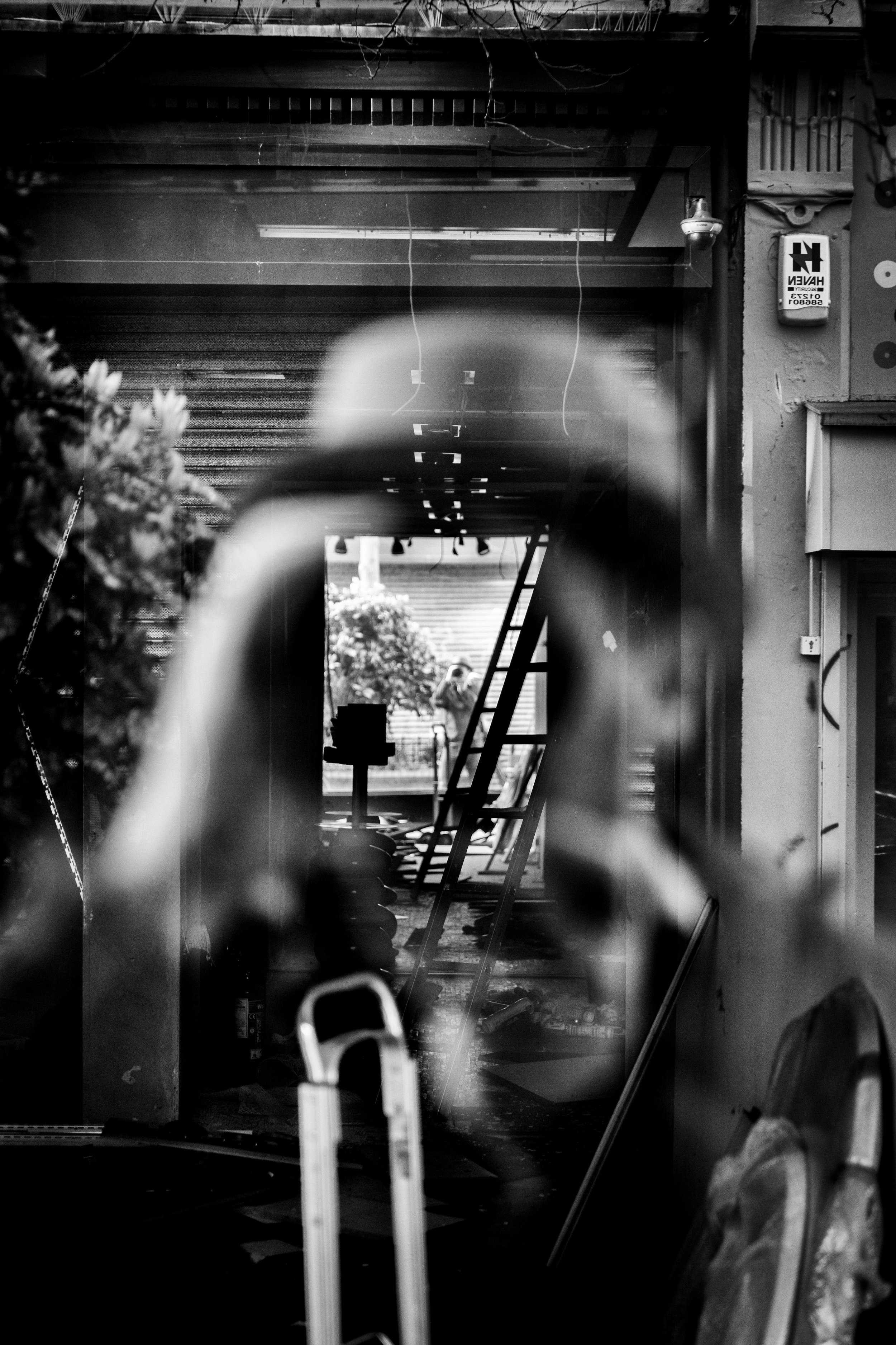 Black and white photo of a garage or workshop space viewed through a blurry, abstract shape in the foreground, with a ladder, workbench, and a person in the background.