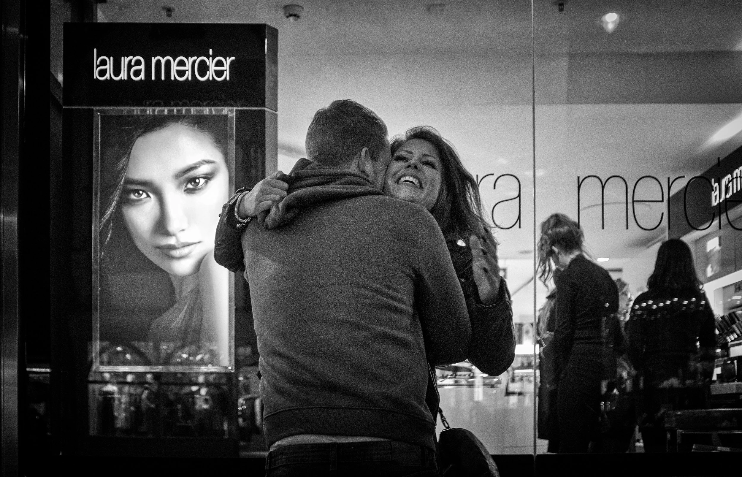 A man and woman hugging and smiling inside a store, with a large advertisement for Laura Mercier cosmetics and other shoppers in the background.