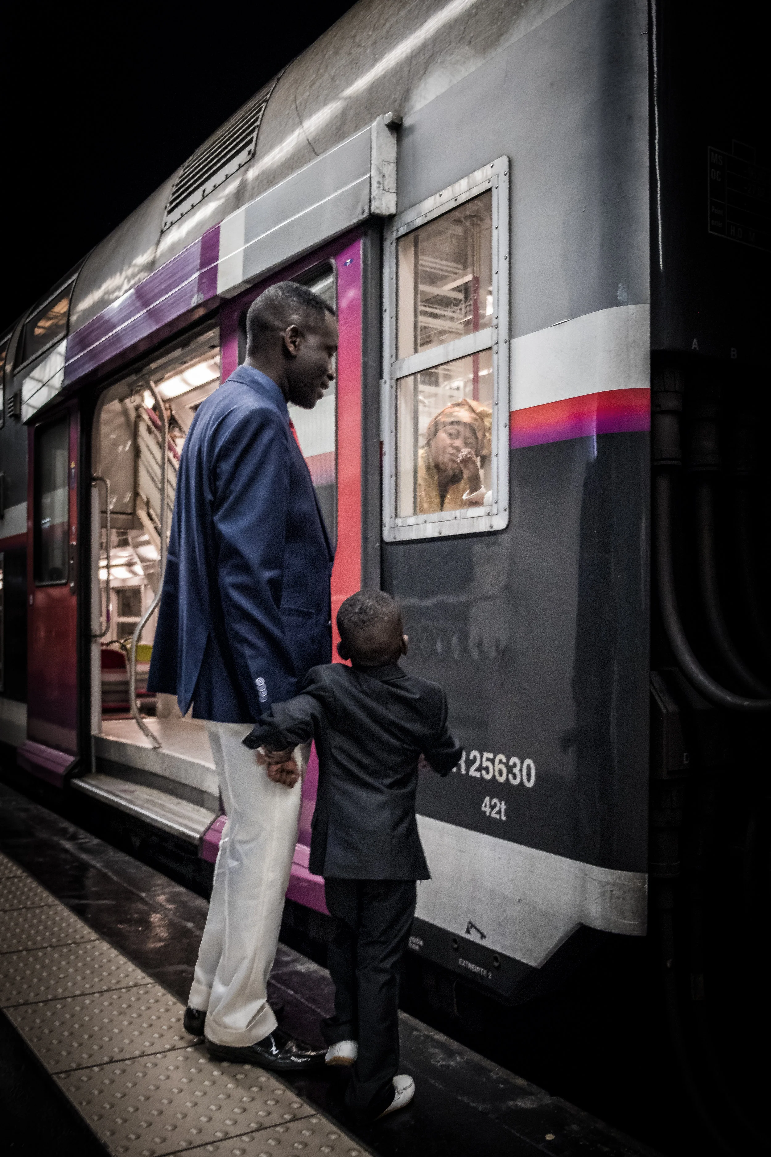 A man and a young boy in suits are standing on a train platform, holding hands, as they look inside the train window where a woman is visible looking back at them.