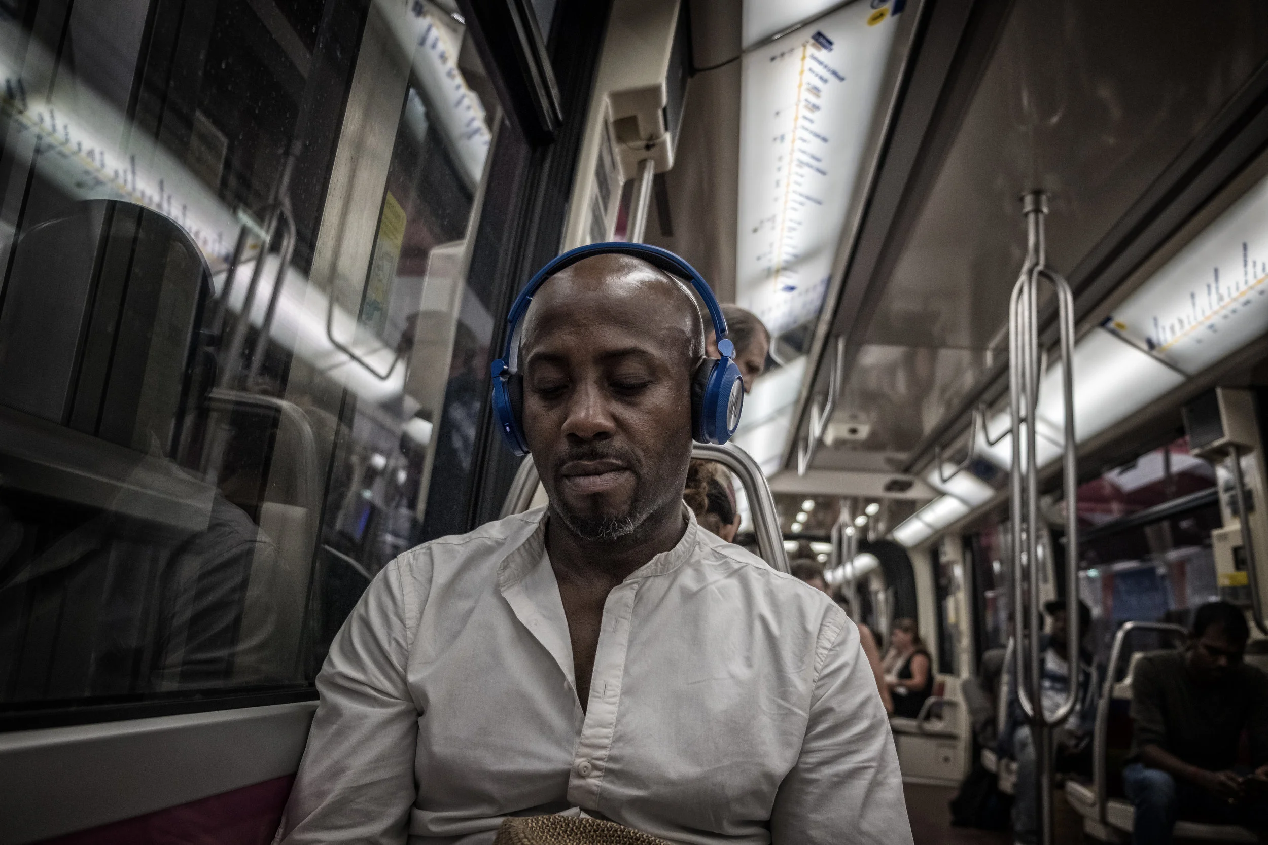A man with a shaved head and beard, wearing a white shirt and blue headphones, sitting on a public transportation vehicle