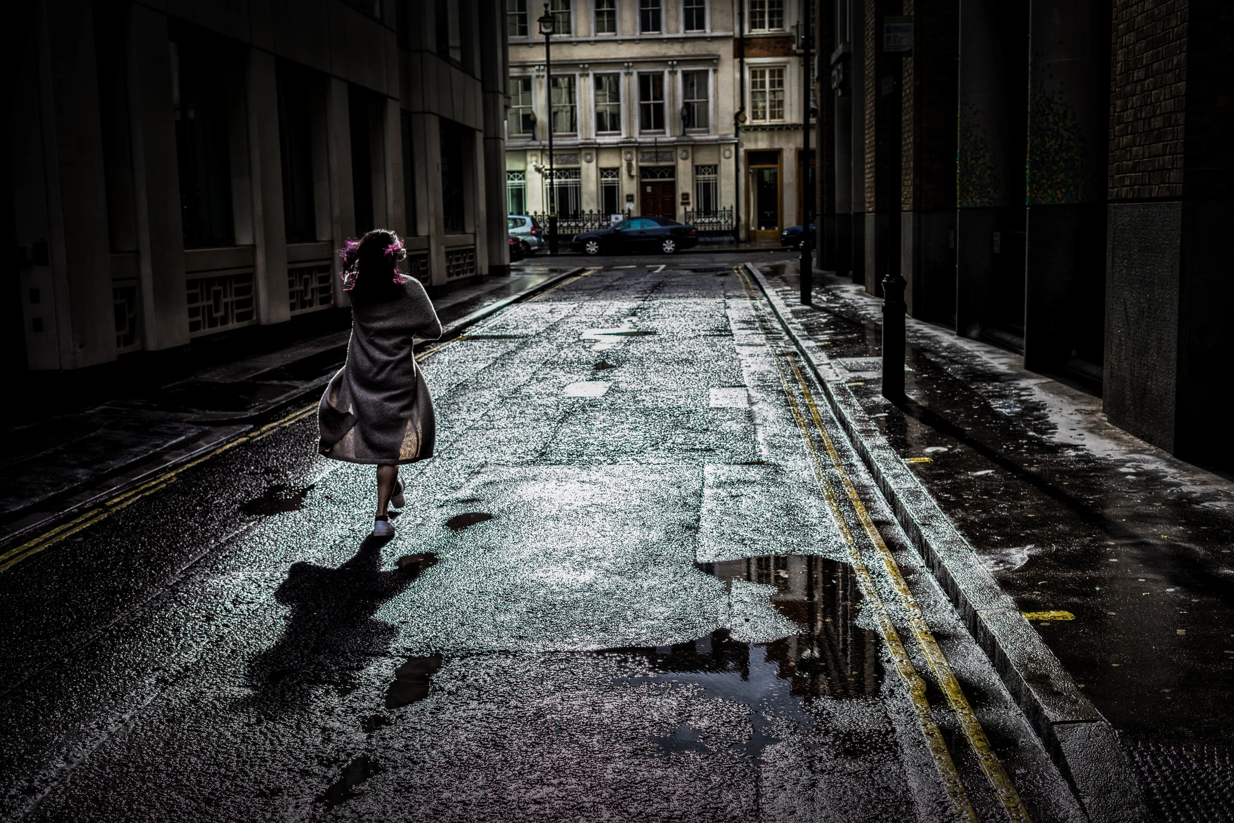 A person with purple hair walking down a wet city street with puddles, surrounded by tall buildings, on a sunny day.