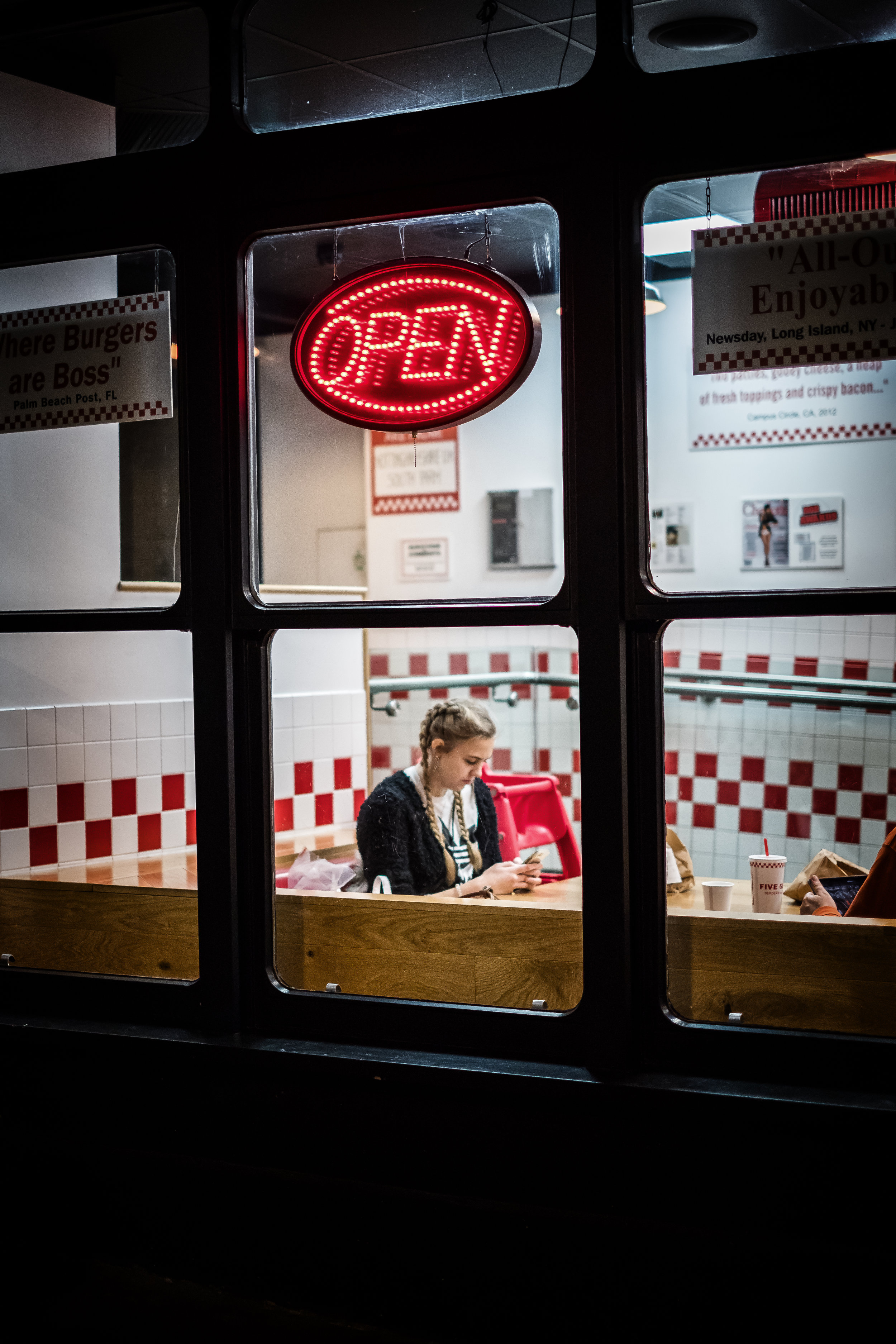 Inside a fast-food restaurant seen through a window, a blonde girl with braids is sitting at a wooden table, looking at her phone. There is a red number 5 cup on the table, paper bags, and a red high chair in the background.