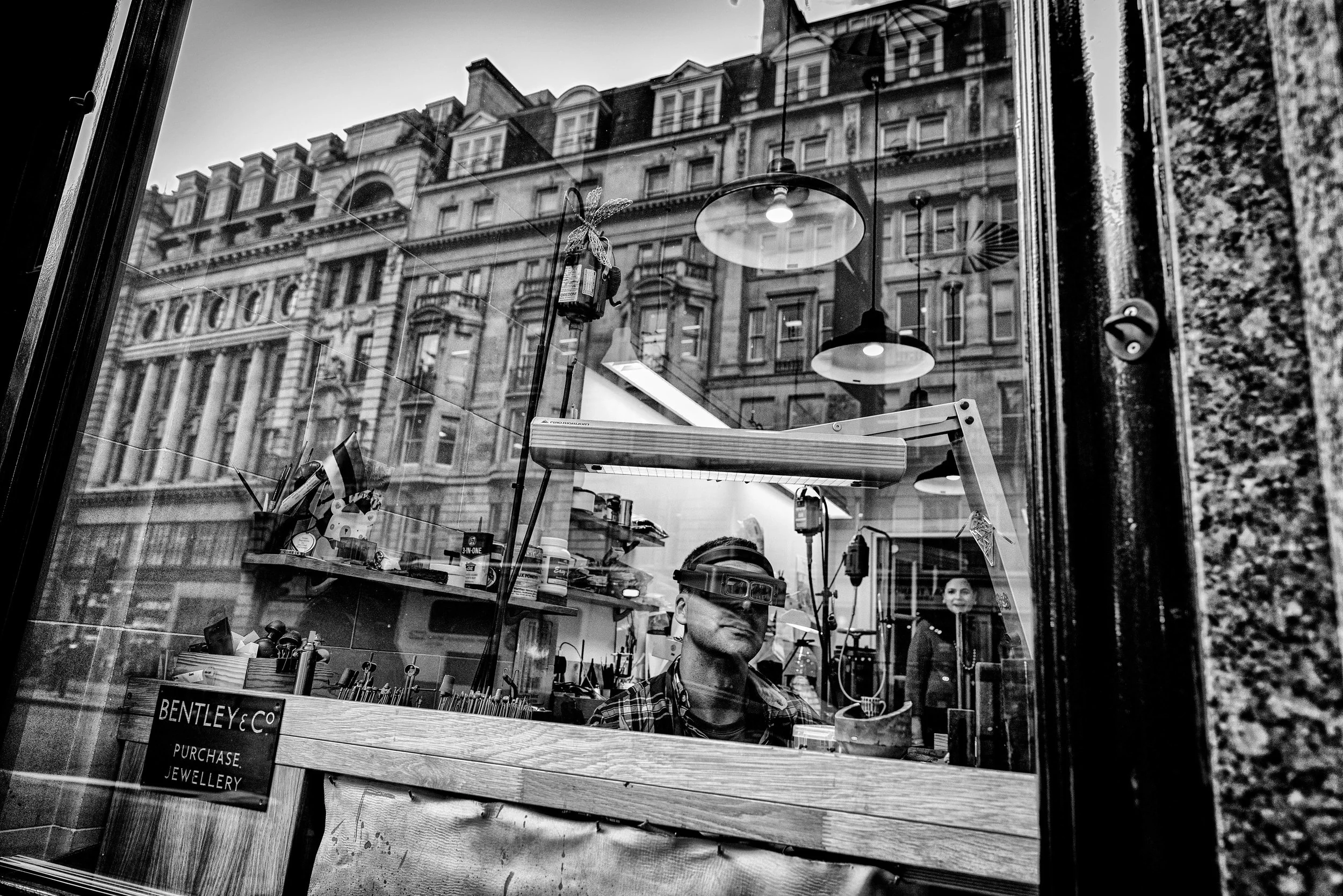 Black and white photo of a jewelry store window, with a worker using jeweler's goggles inside. The reflection of historic building facades is visible in the glass.