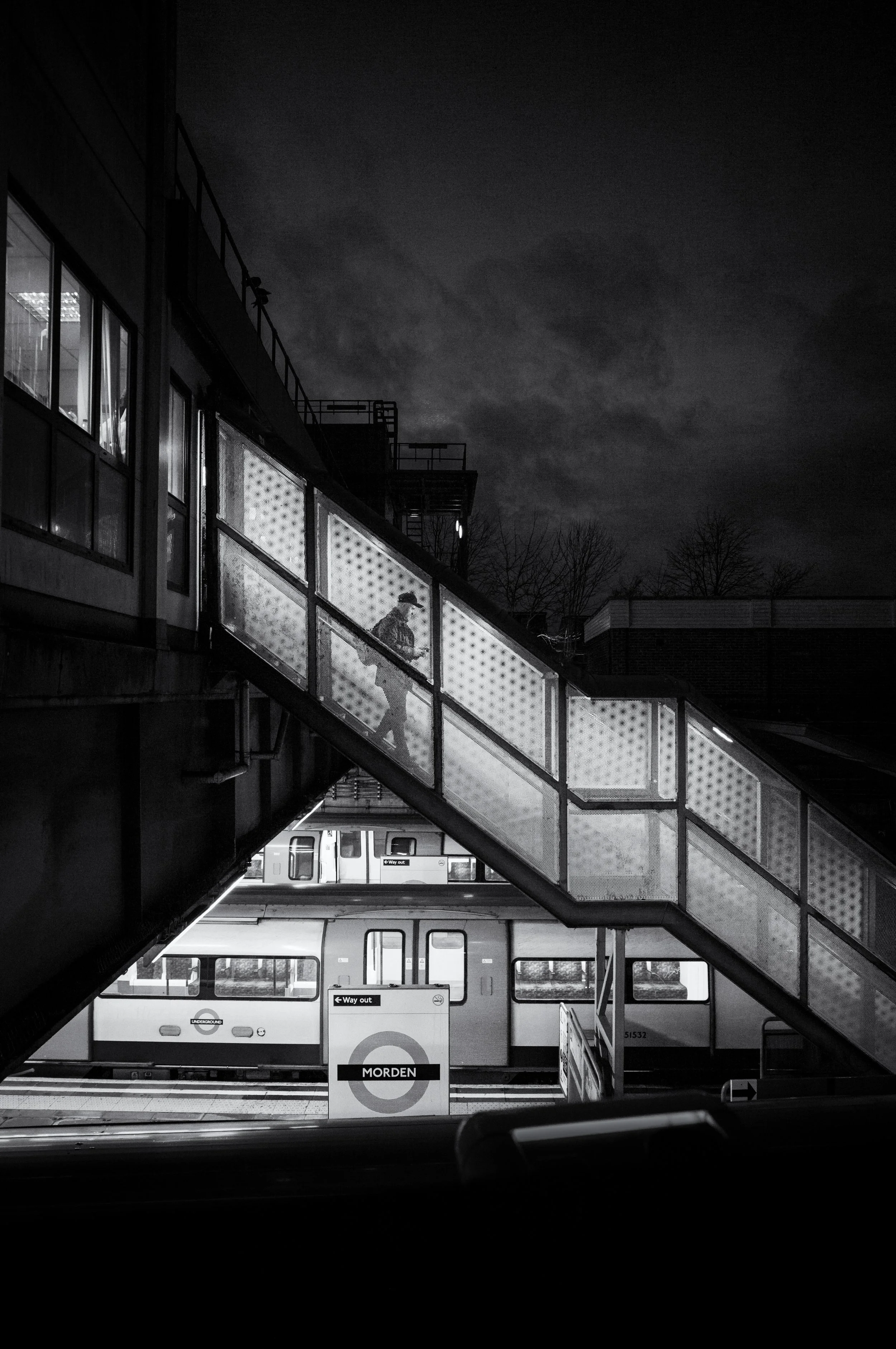Nighttime scene of a train and an escalator at Morden station, with a person walking on the escalator. The sky is cloudy and dark.