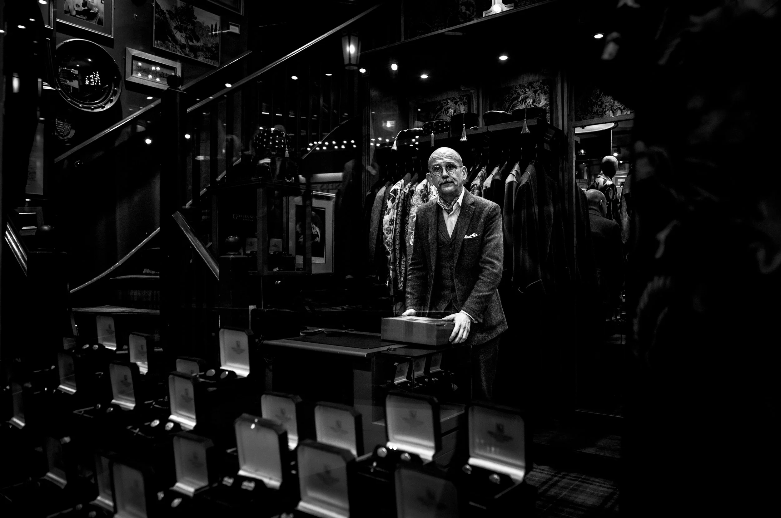 A man with glasses and a beard in a suit appears to be at a jewelry store or a luxury shop, standing behind a counter with jewelry boxes, with clothing racks and framed pictures in the background.