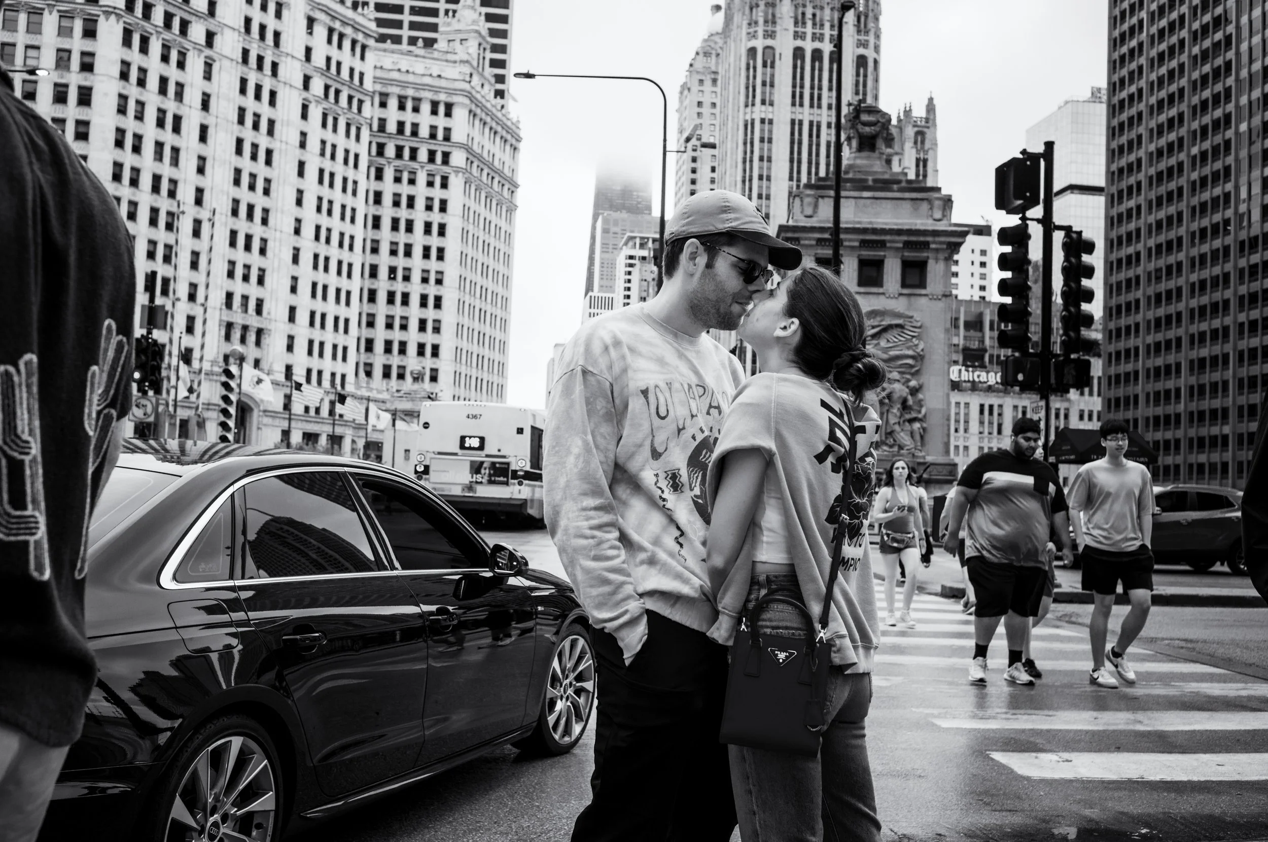 A couple sharing a kiss in a Chicago street, with tall buildings, cars, and pedestrians in the background.