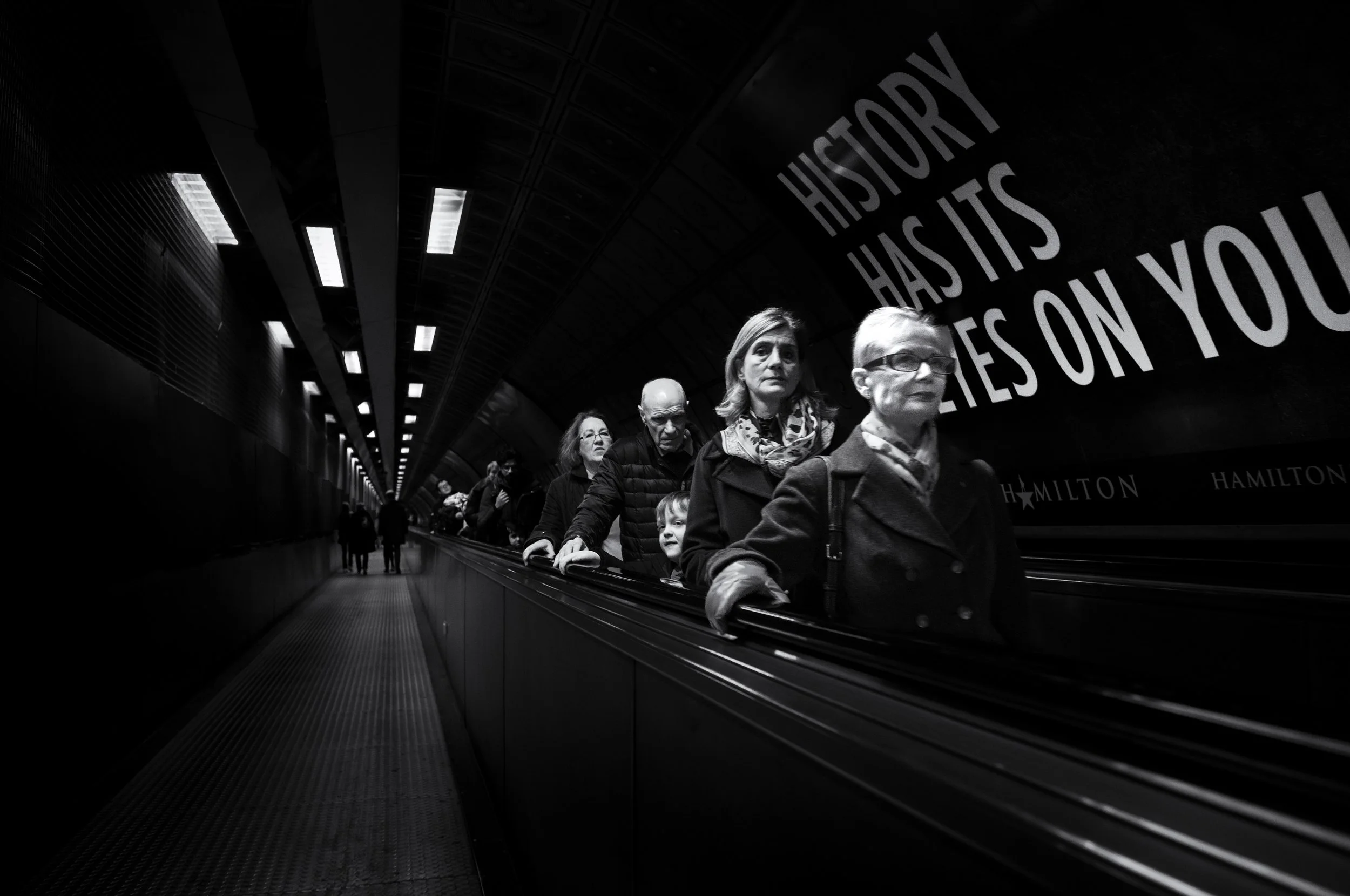 People riding escalator in underground station with large quote on wall reading 'History has its eyes on you'.