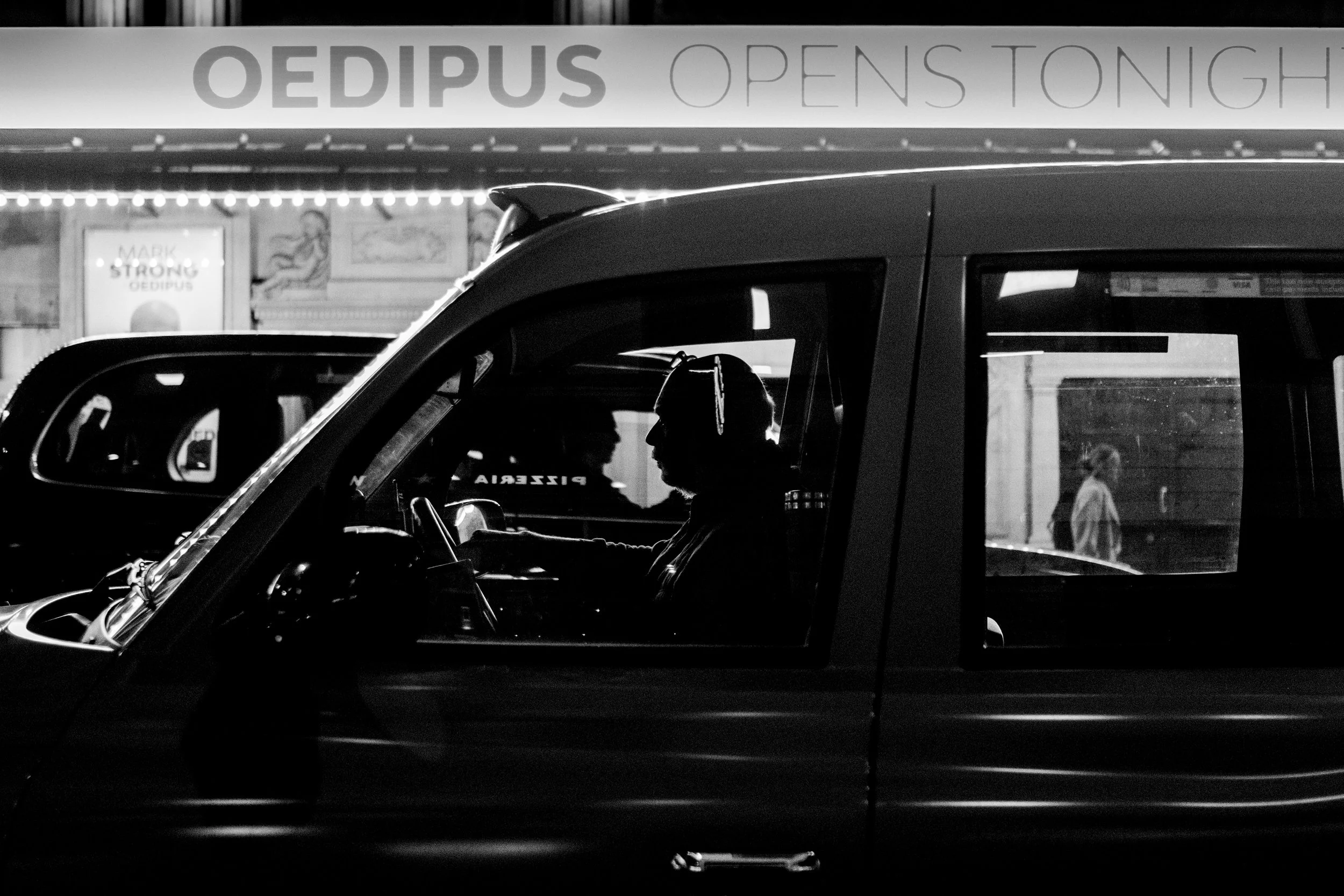 A black and white photo showing a person driving a taxi in an urban area at night. The silhouette of the driver is visible through the window, with a sign in the background reading 'OEDIPUS OPEN TONIGHT' and other pedestrians walking by.