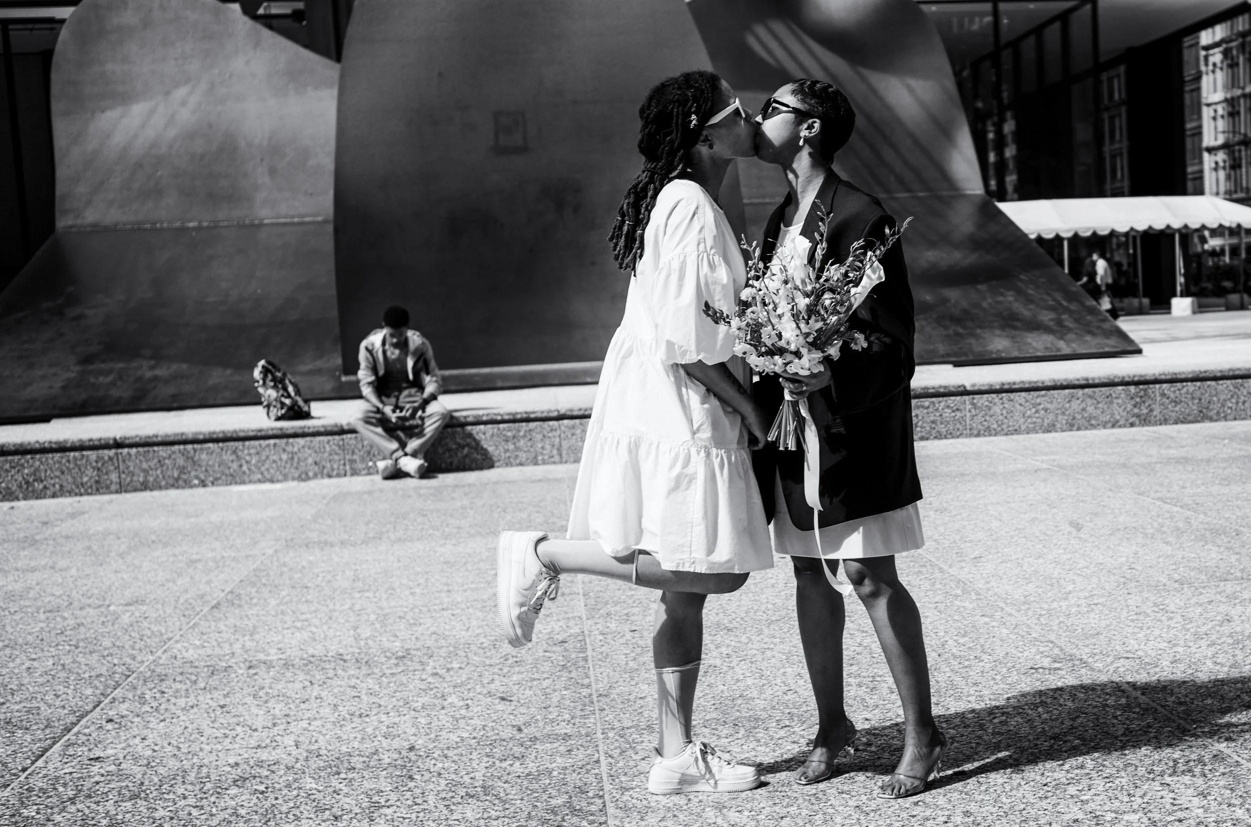 Two women sharing a kiss in an urban park, one holding a bouquet of flowers. A person sits on the ground in the background.