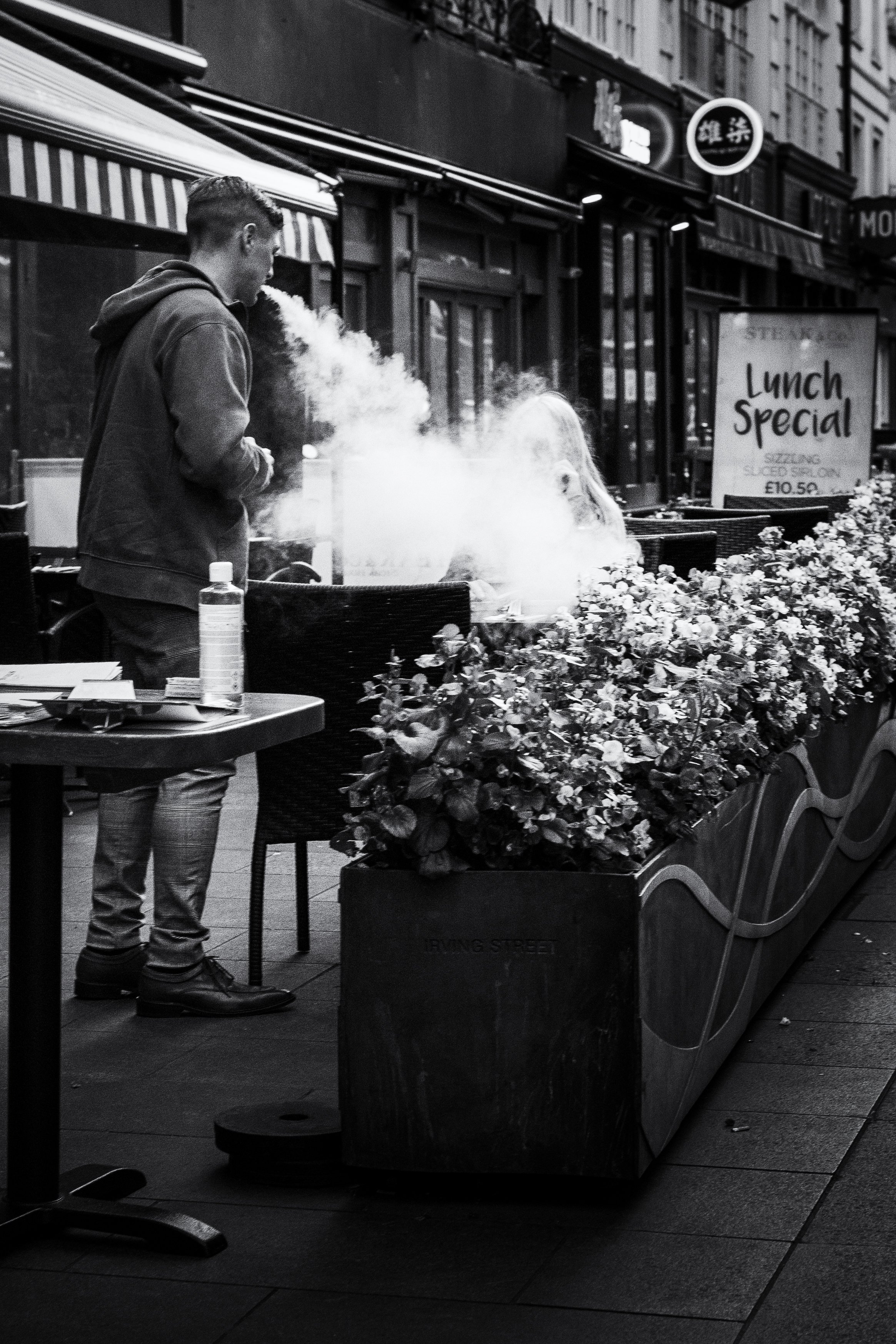 A young man in a hoodie and pants standing outside a restaurant, holding a smoking item, with a woman sitting at the table nearby. The restaurant has large windows, a sign that says 'Lunch Special,' and a flower planter in the foreground.