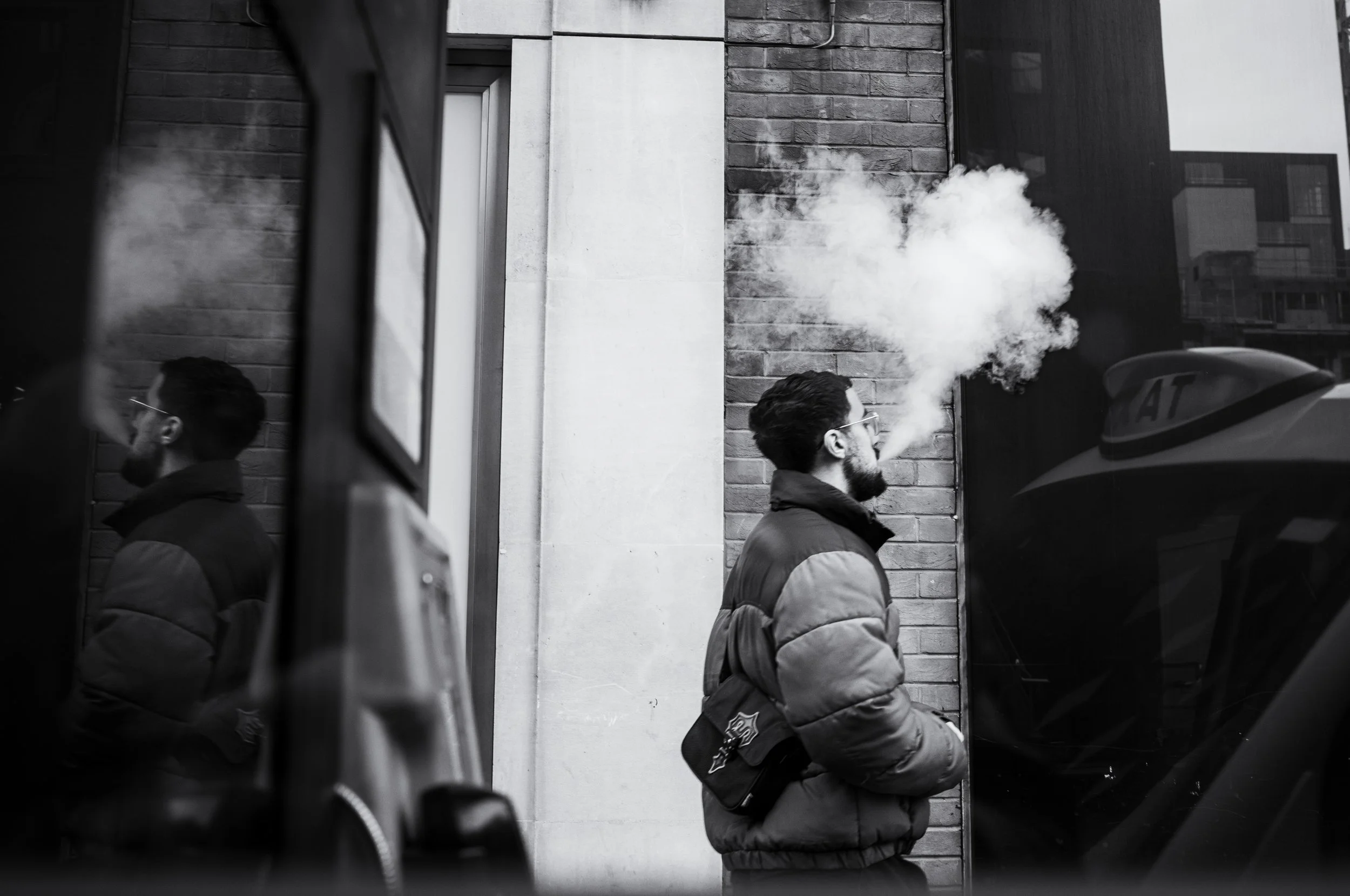 A man with glasses and a beard in a puffy jacket is exhaling vapor or smoke against a brick wall in an urban setting, reflected in a window.