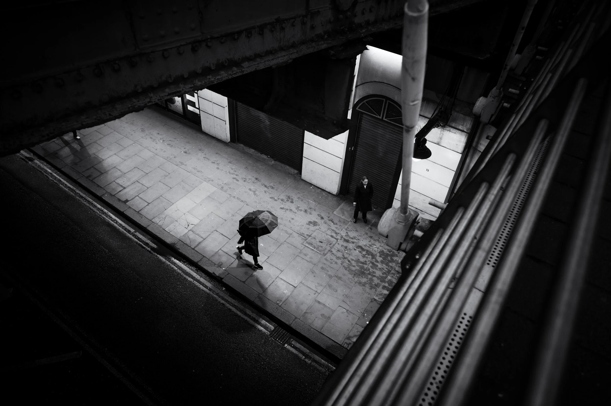 Black and white photo of a city street viewed from above, showing two pedestrians walking under umbrellas, one near a building with closed shutters, the other on the sidewalk.