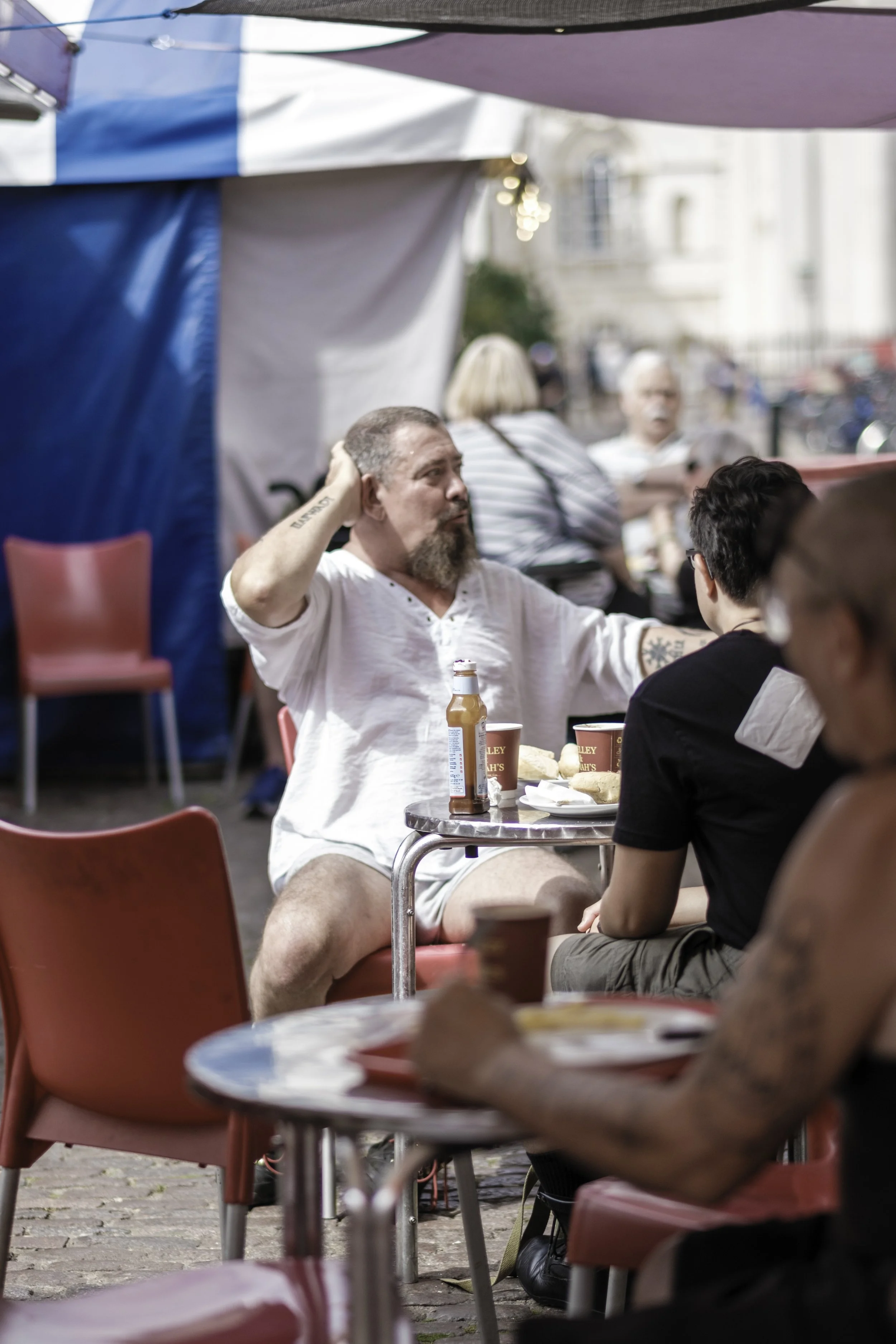 People sitting at outdoor tables, eating, drinking, and conversing, under tents with a background of blurred buildings and street scene.