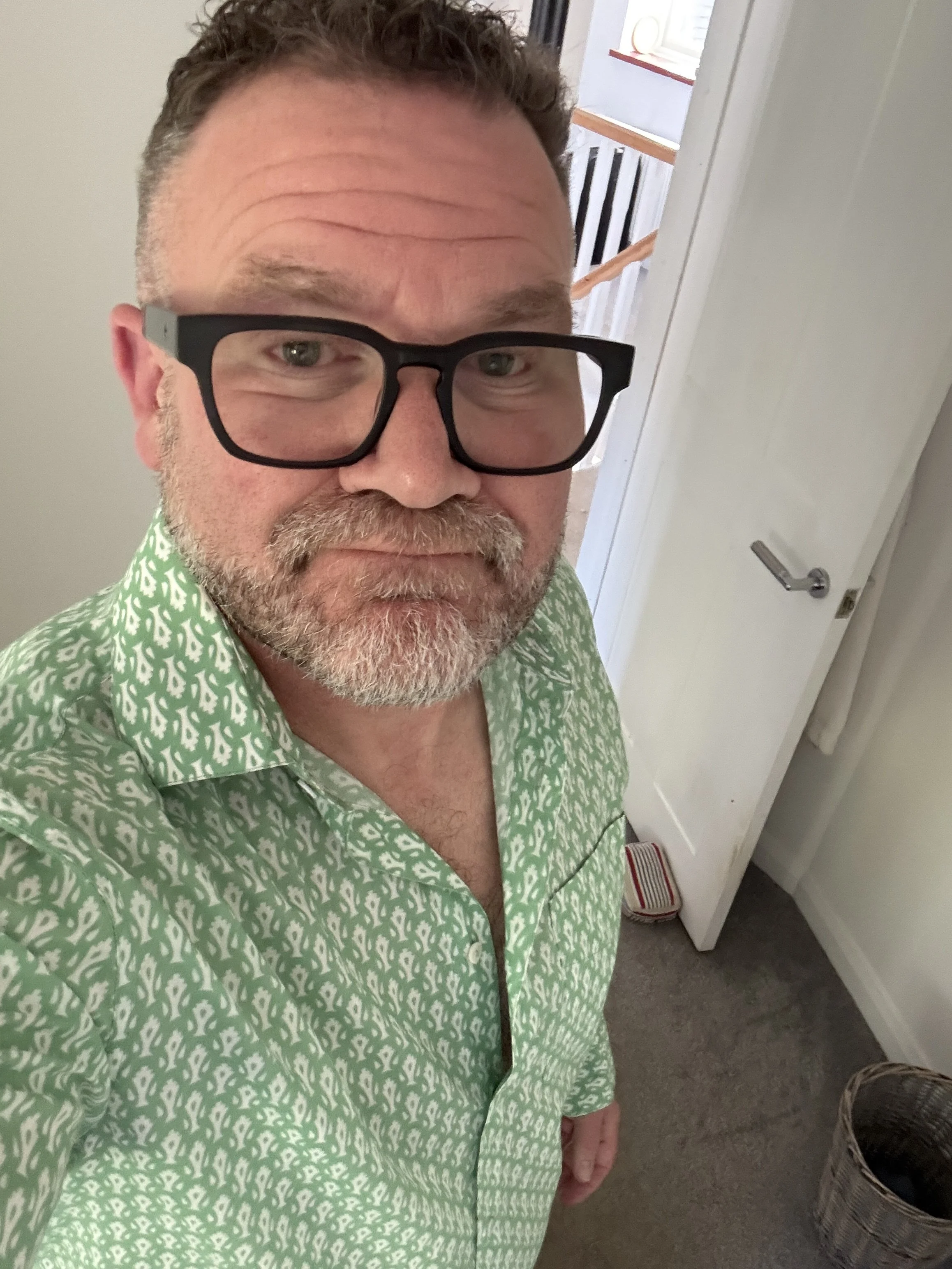 A man with glasses and a beard taking a selfie indoors, wearing a green patterned shirt, standing near a door.