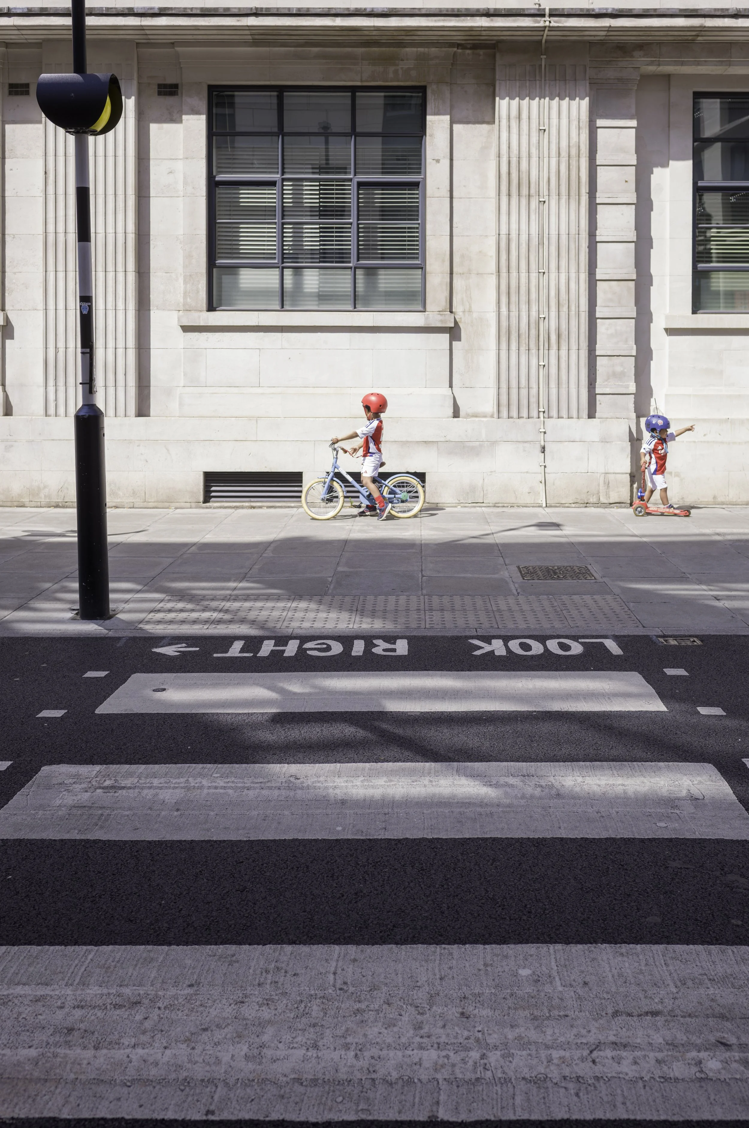 Two children in helmets riding bikes and scooters across a city street with a pedestrian crosswalk, with a building in the background and a black pole on the left.