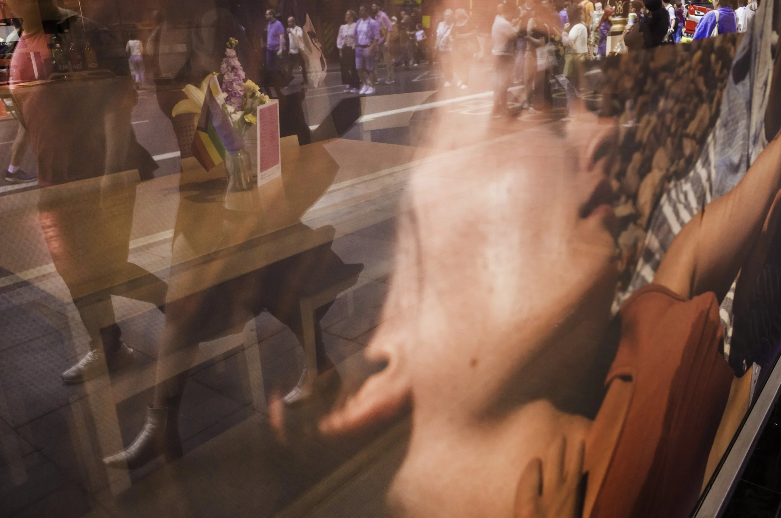 Reflection of a person with glasses and freckles looking at a window display, with a street scene of people walking outside visible through the window.