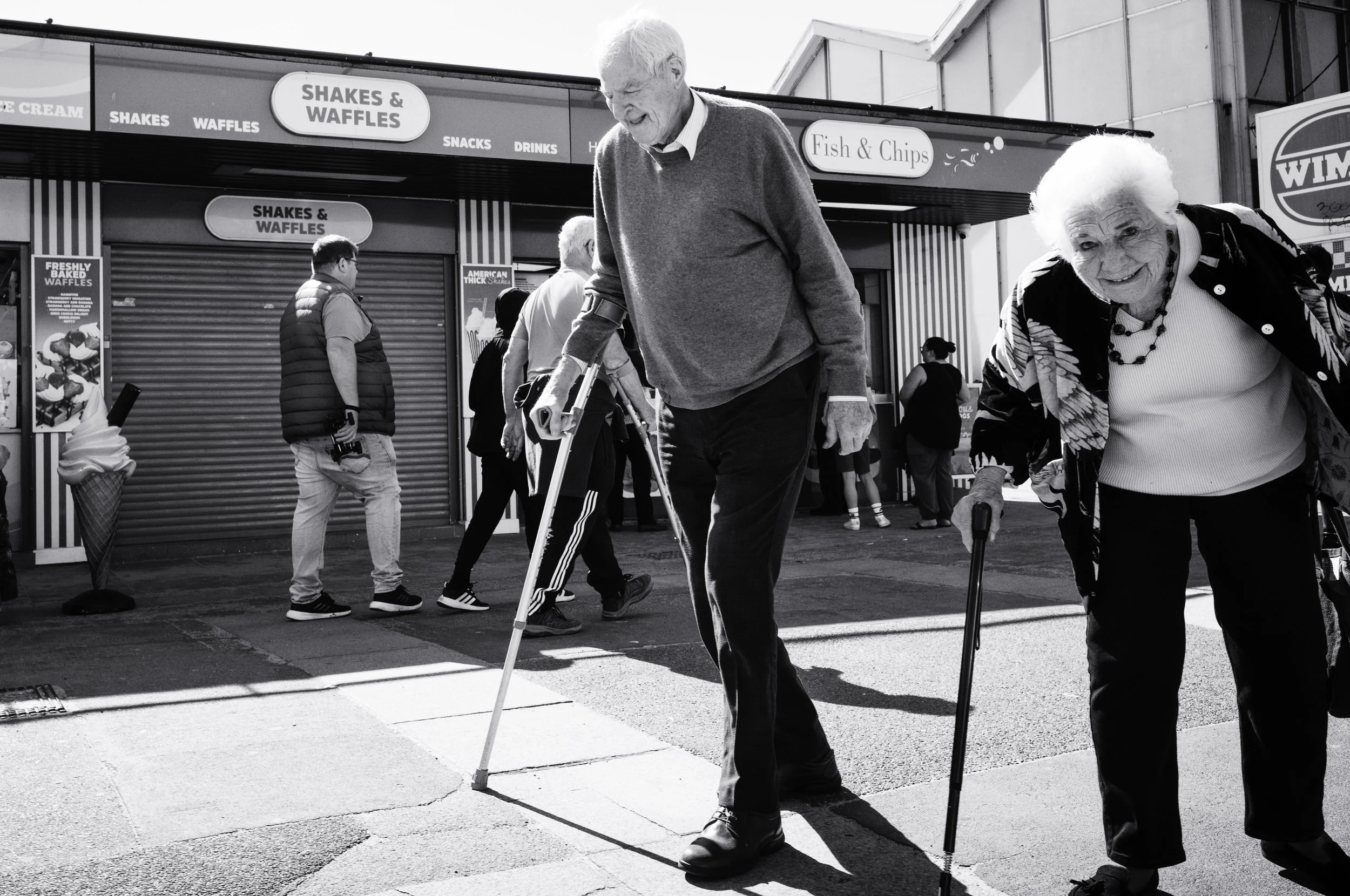 Two elderly people with canes walking in front of a food stand at an outdoor fair or market. The stand sells shakes, waffles, fish, and chips, and has a sign for ice cream and ice cream cones.