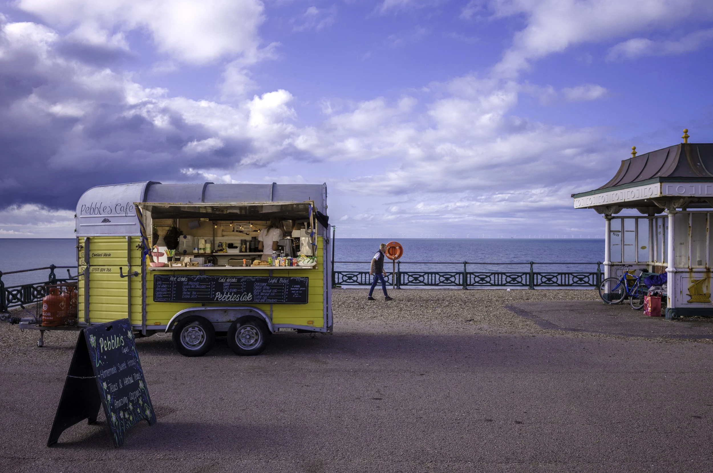 A yellow food truck labeled 'Pebbles Cafe' parked on a promenade overlooking the ocean, with a chalkboard menu, a person walking past, and a nearby ornate kiosk with bicycles leaning against it.