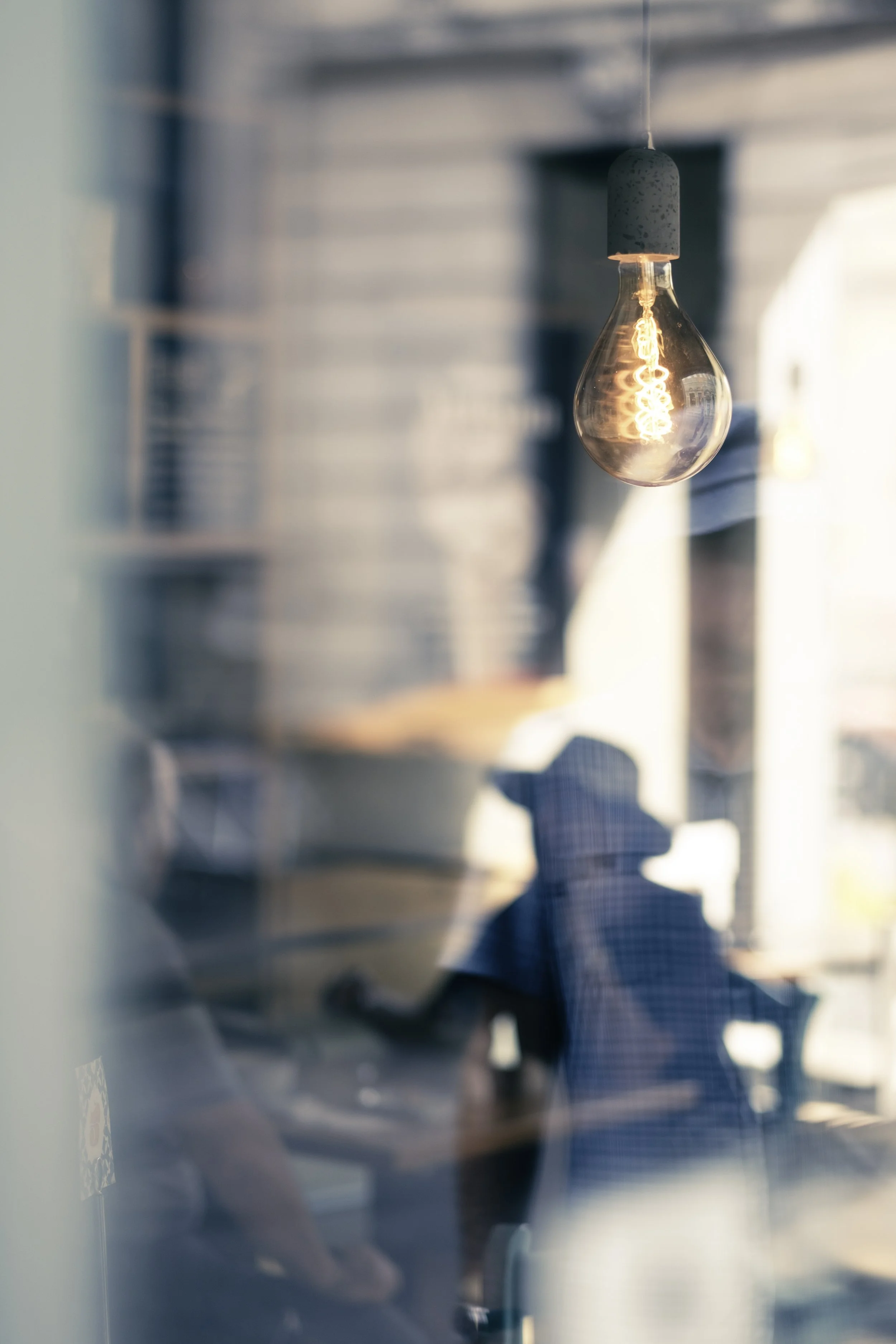A hanging light bulb with a visible filament, blurred background showing people and interior space.