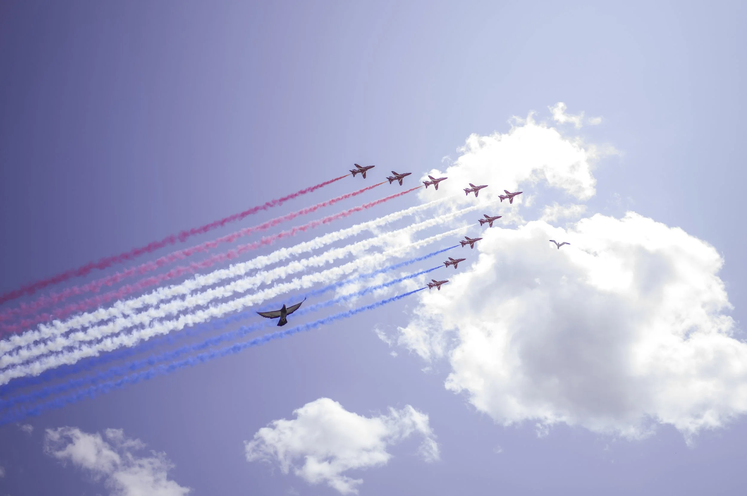 A formation of nine jets flying in a curve in the sky, leaving colored smoke trails in red, white, and blue, with clouds and a blue sky in the background.