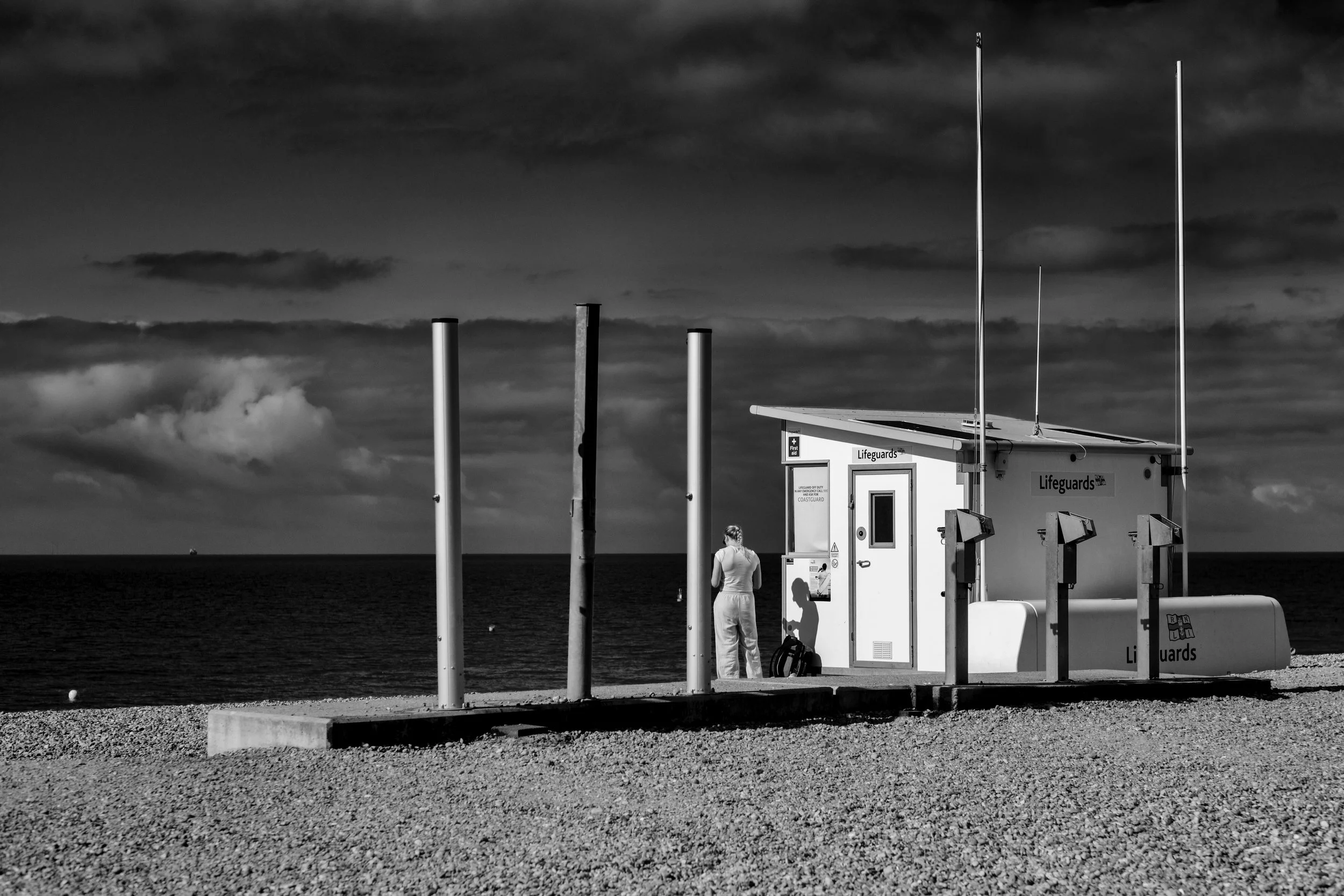 A person standing outside a lifeguard station on a beach, with the ocean in the background, black and white photo.