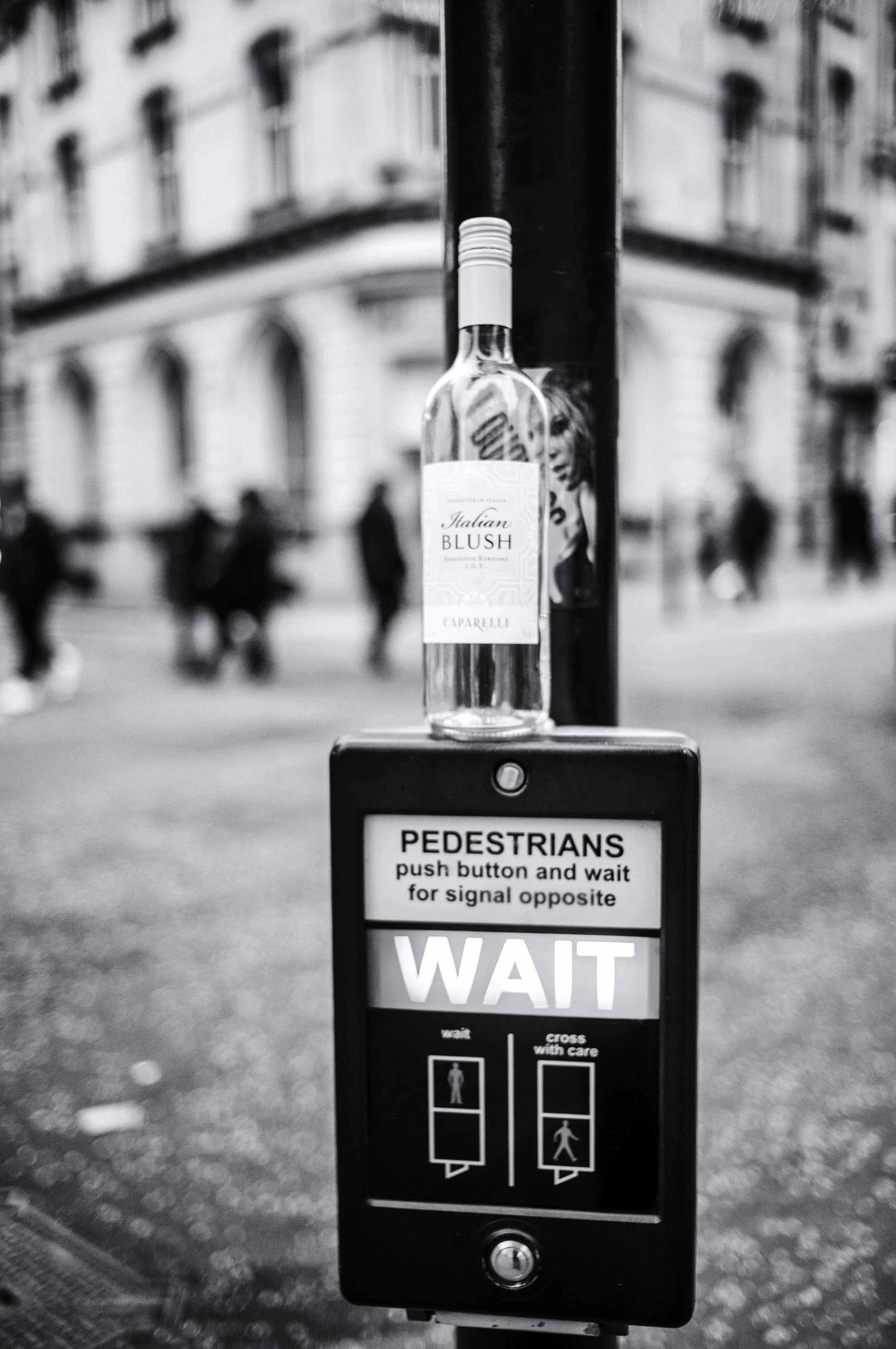 A pedestrian crossing button station on a city street with a bottle of Italian Blush wine placed on top. The sign instructs pedestrians to push button and wait for signal opposite, with callouts to wait or cross with care.