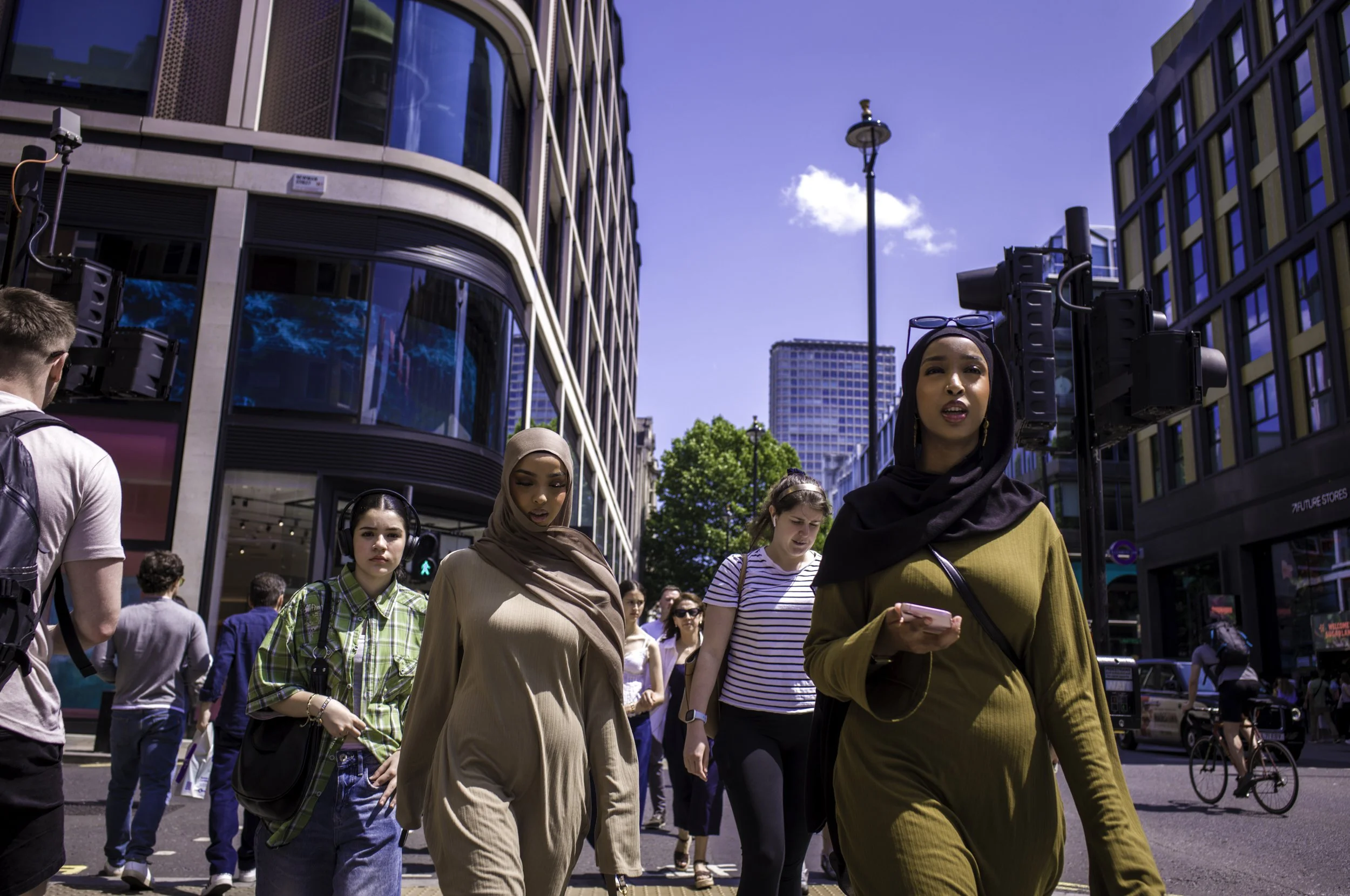 Crowd of diverse pedestrians crossing a city street on a sunny day with modern buildings in the background.