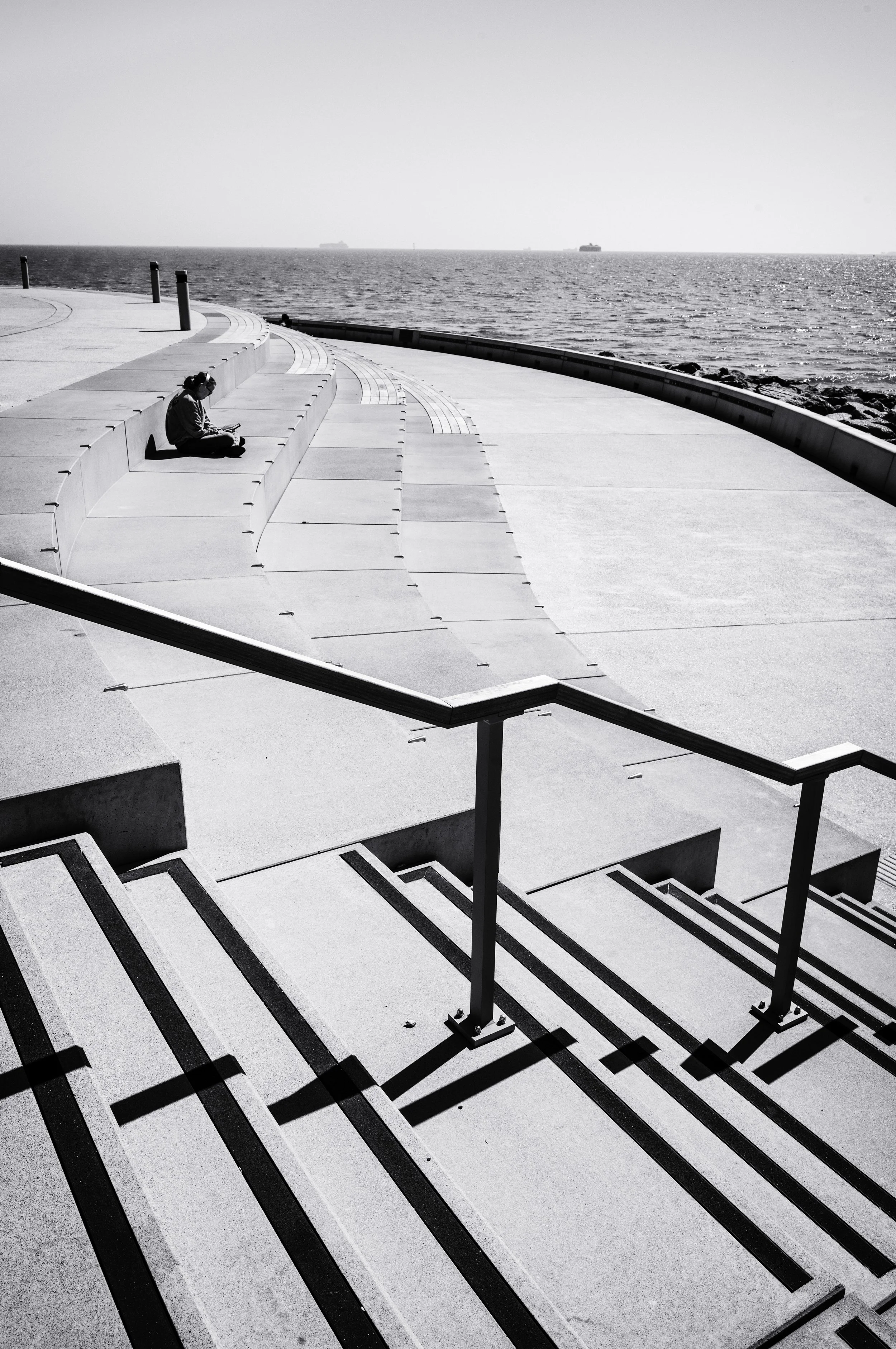 A person sitting on concrete steps by the water, reading a book, in black and white.