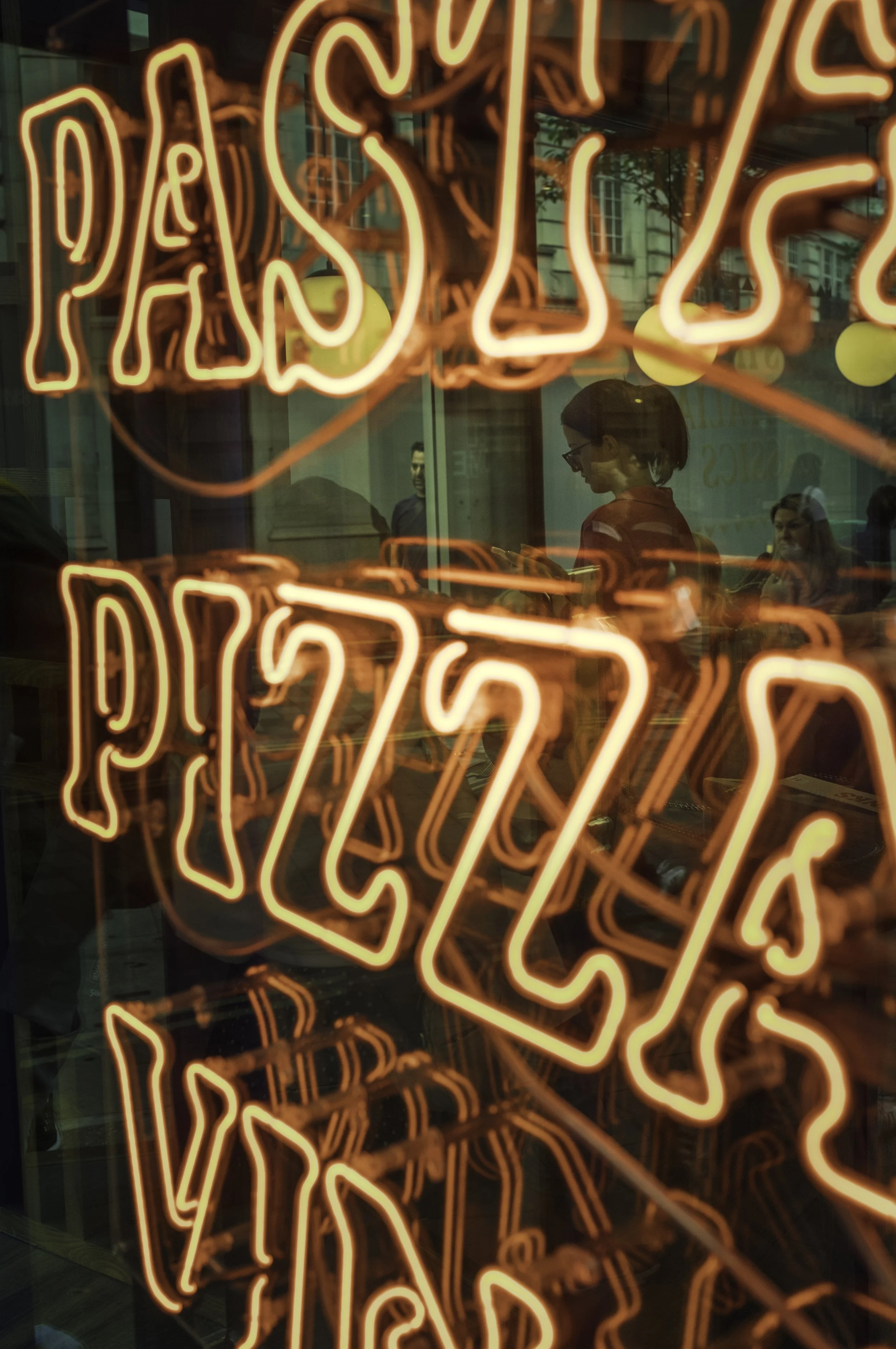 Bright orange neon sign that reads 'Pastries' in a bakery storefront, with reflections of customers and city buildings visible in the glass.
