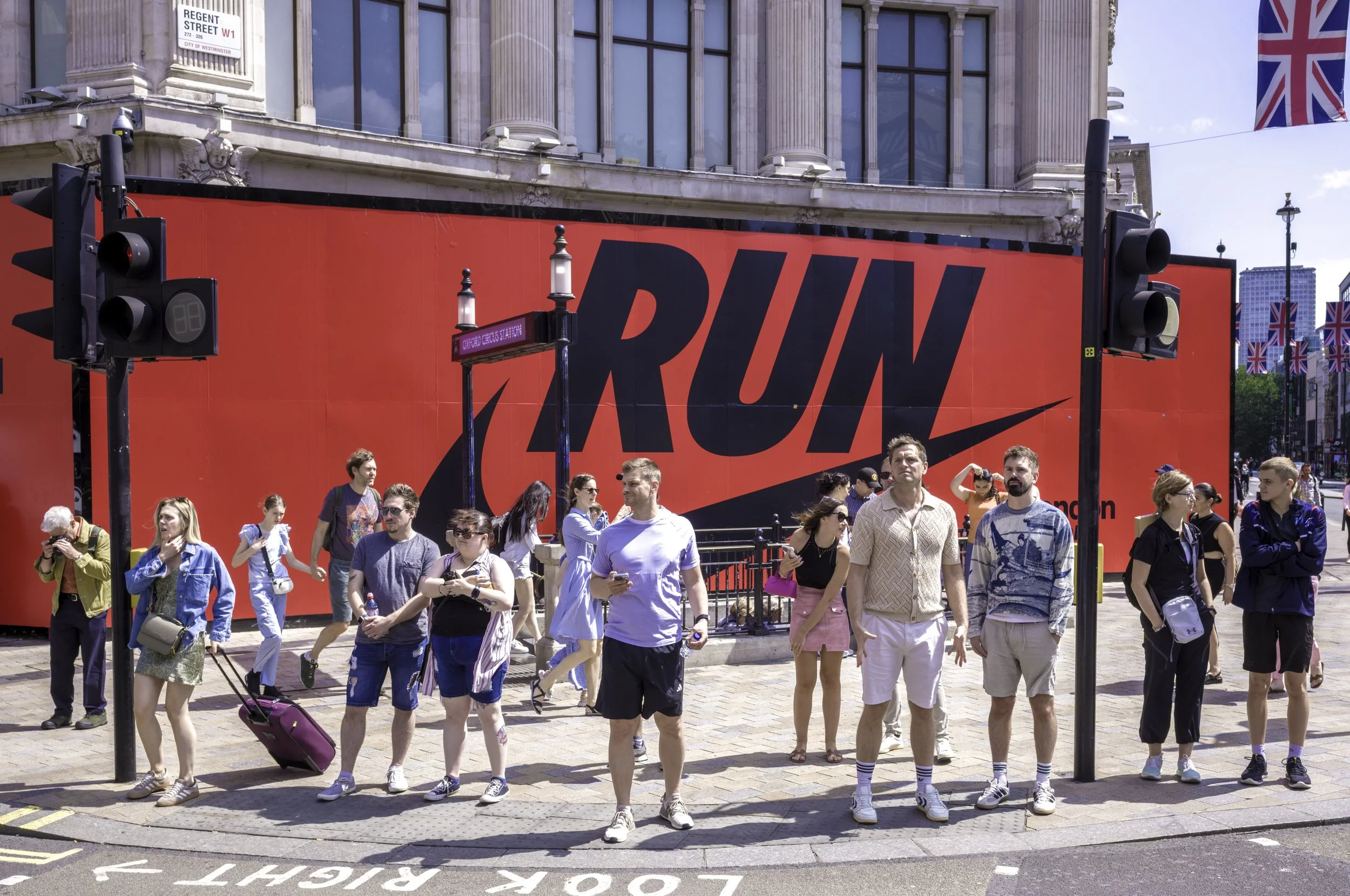 Crowd of people waiting at a crosswalk in front of a large red Nike billboard with the word 'RUN' in bold black letters, on a city street with British flags hanging.