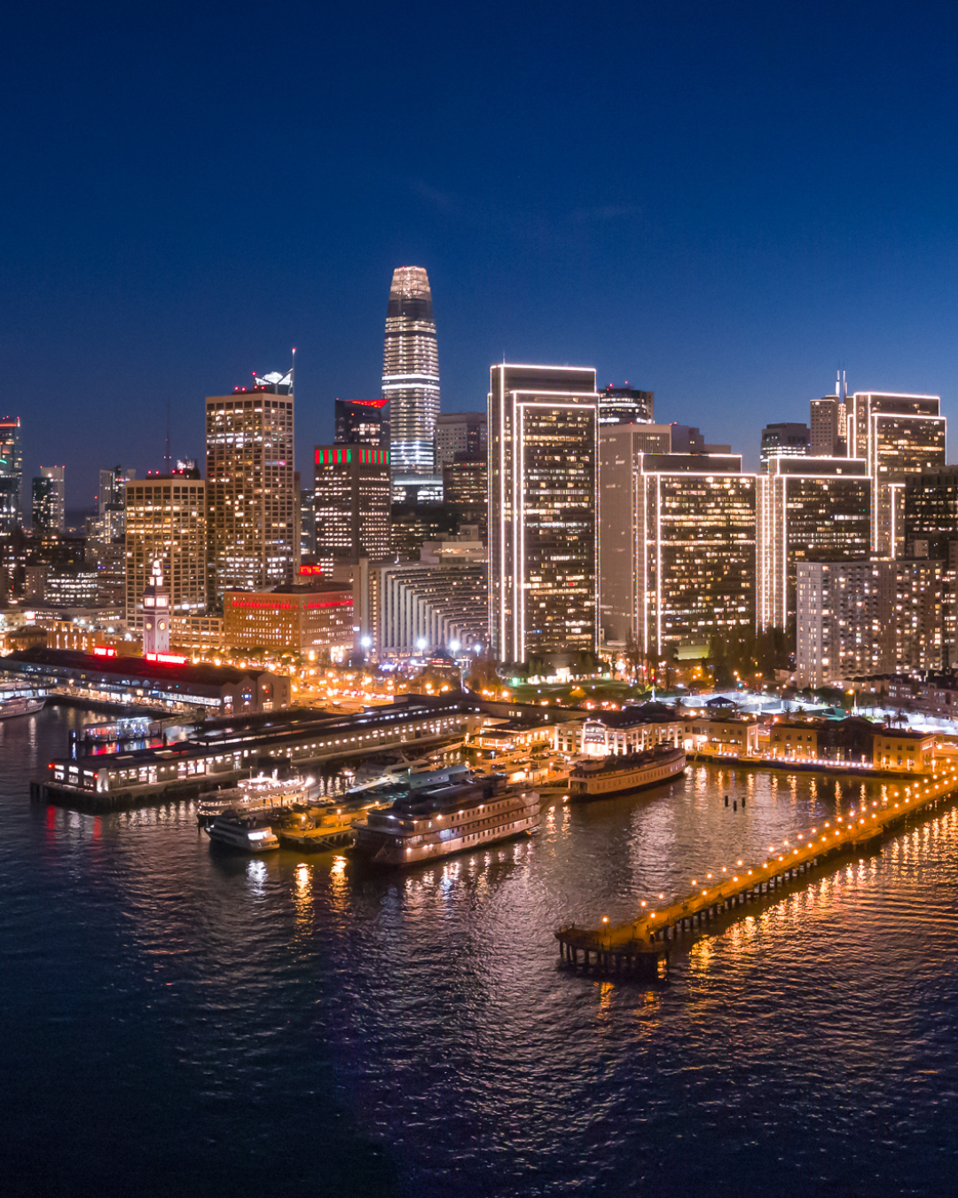 Night view of a city skyline with tall illuminated buildings and a waterfront with boats and a pier.