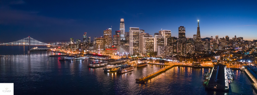 Nighttime aerial view of the San Francisco skyline with illuminated skyscrapers, the Bay Bridge on the left, and waterfront piers in the foreground.