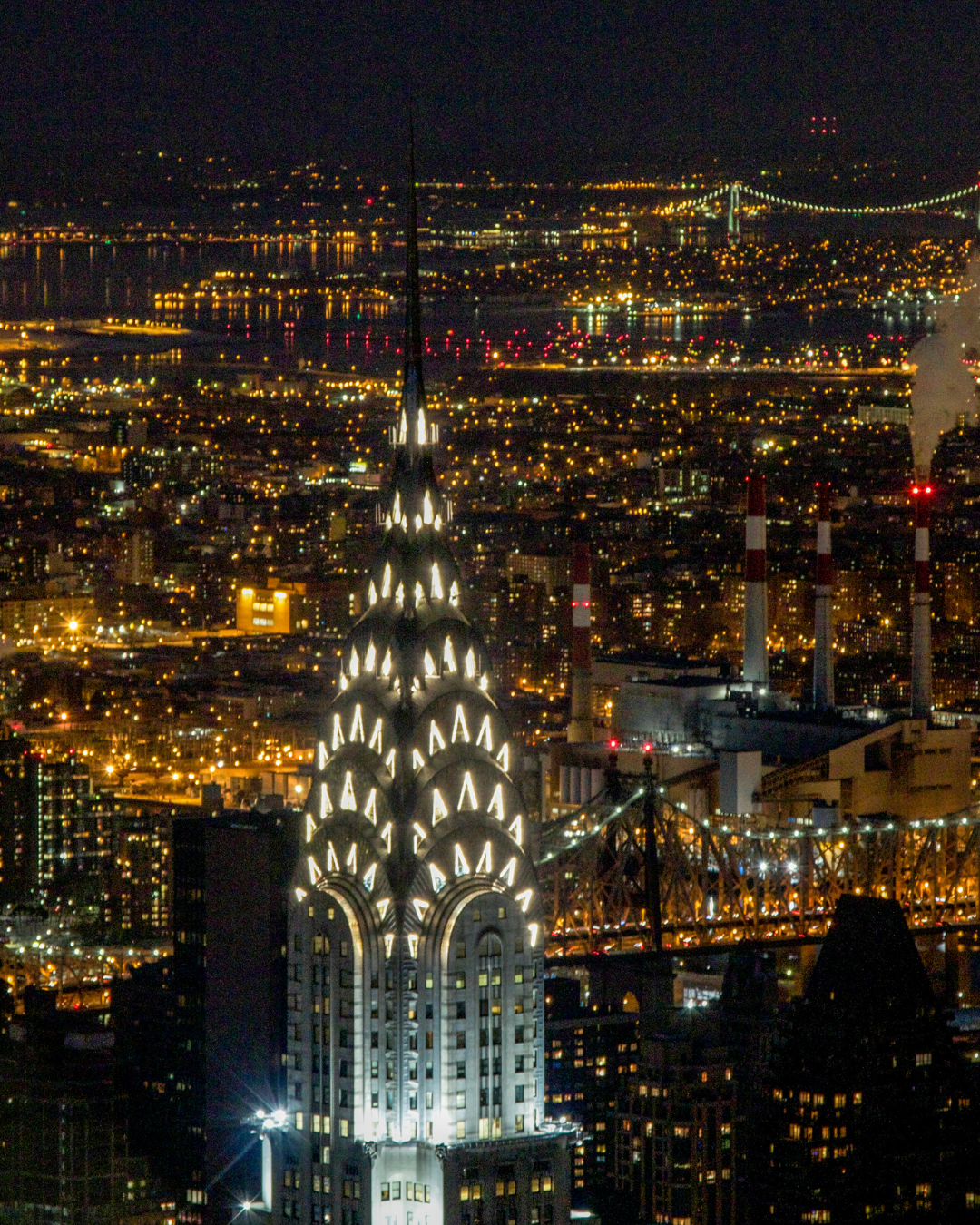 Nighttime aerial view of the New York City skyline, featuring the illuminated Chrysler Building in the foreground, with the Queensboro Bridge and East River in the background.