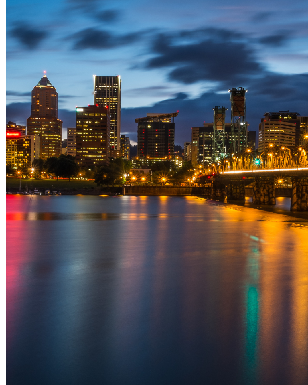 Nighttime city skyline with tall buildings, illuminated windows, and a bridge over water reflecting colorful lights.