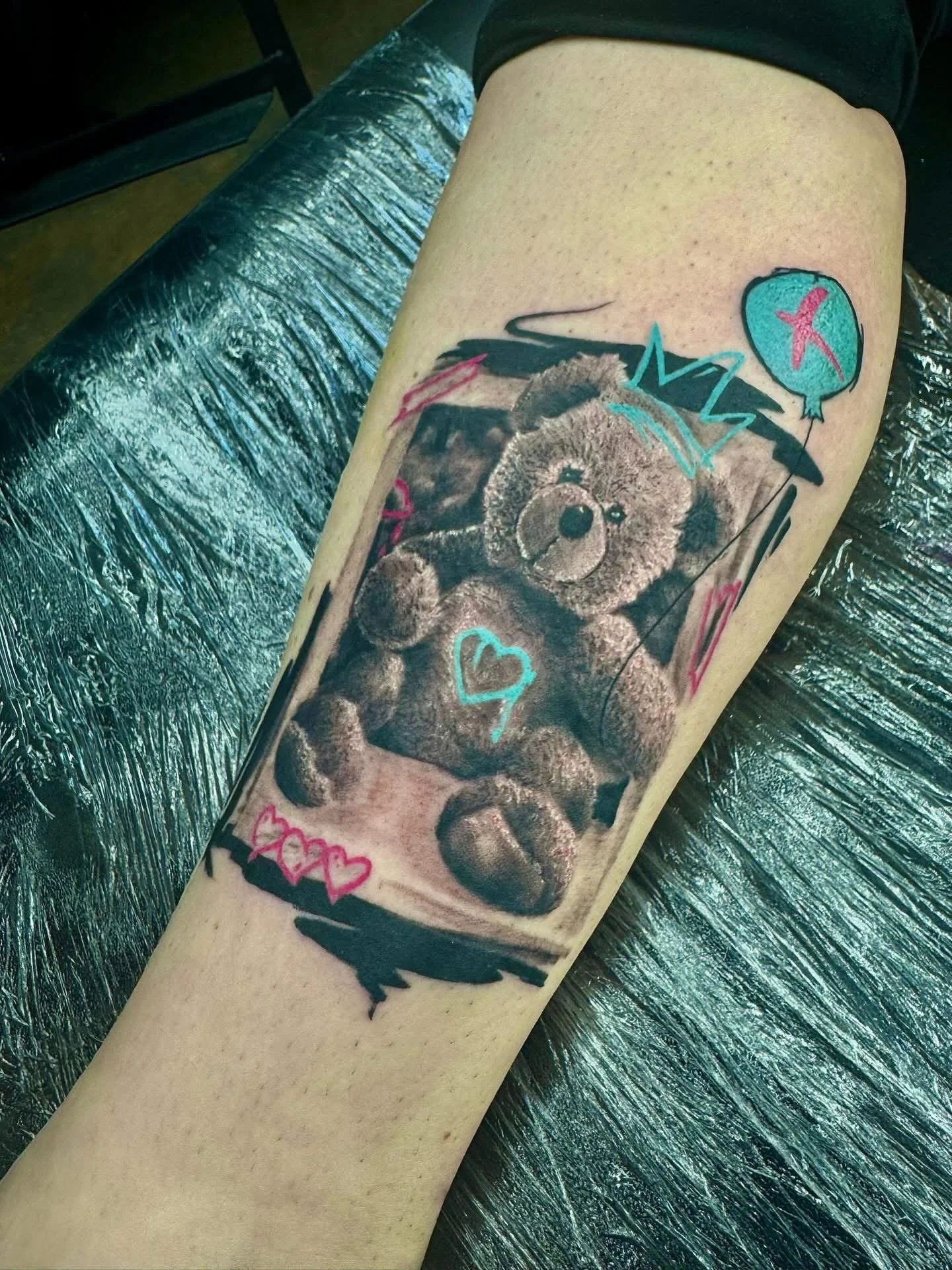 Tattoo of a teddy bear with a neon heart on its chest, wearing a crown, surrounded by colorful doodles including a speech bubble with a starfish, hearts, and abstract lines.