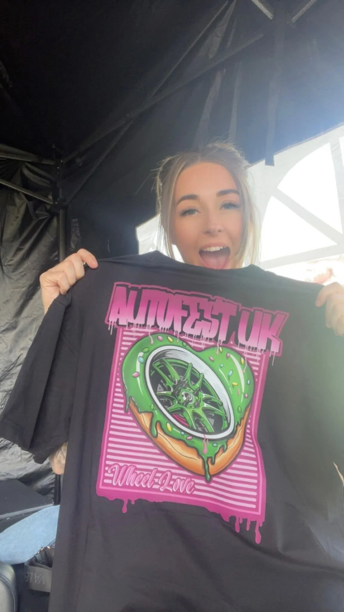 A young woman is holding up a black T-shirt with a colorful graphic design. The shirt features a green slime-covered car wheel with pink and purple accents, and the text 'UK WHEELZ' and 'Wheel-E-Love.'