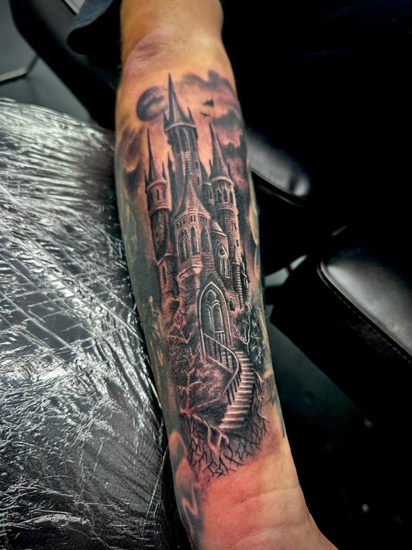Tattoo of a dark, gothic-style castle with multiple towers, steps leading up to it, and tree roots underneath on a person's forearm.