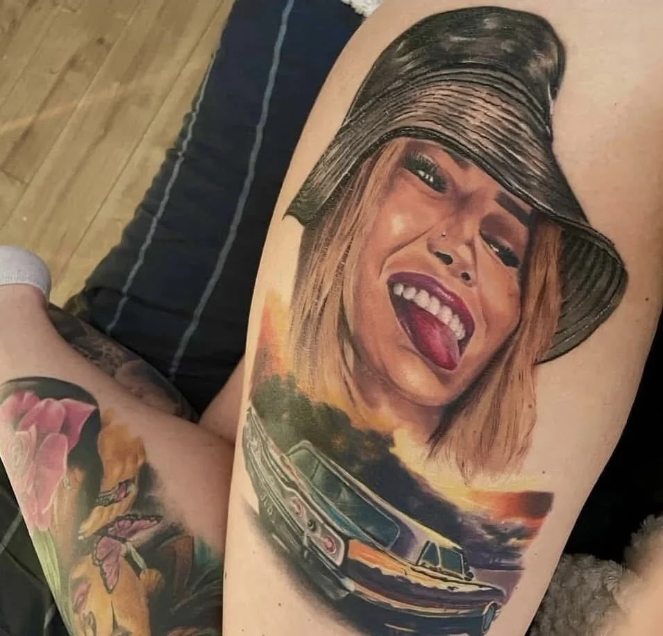 Tattoo of a smiling woman with a nose piercing, wearing a bucket hat and sticking out her tongue, with a car and sunset in the background.