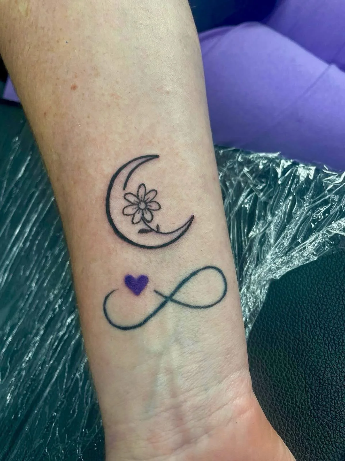 Tattoo of a crescent moon with a flower inside, a purple heart, and an infinity symbol on a person's forearm.